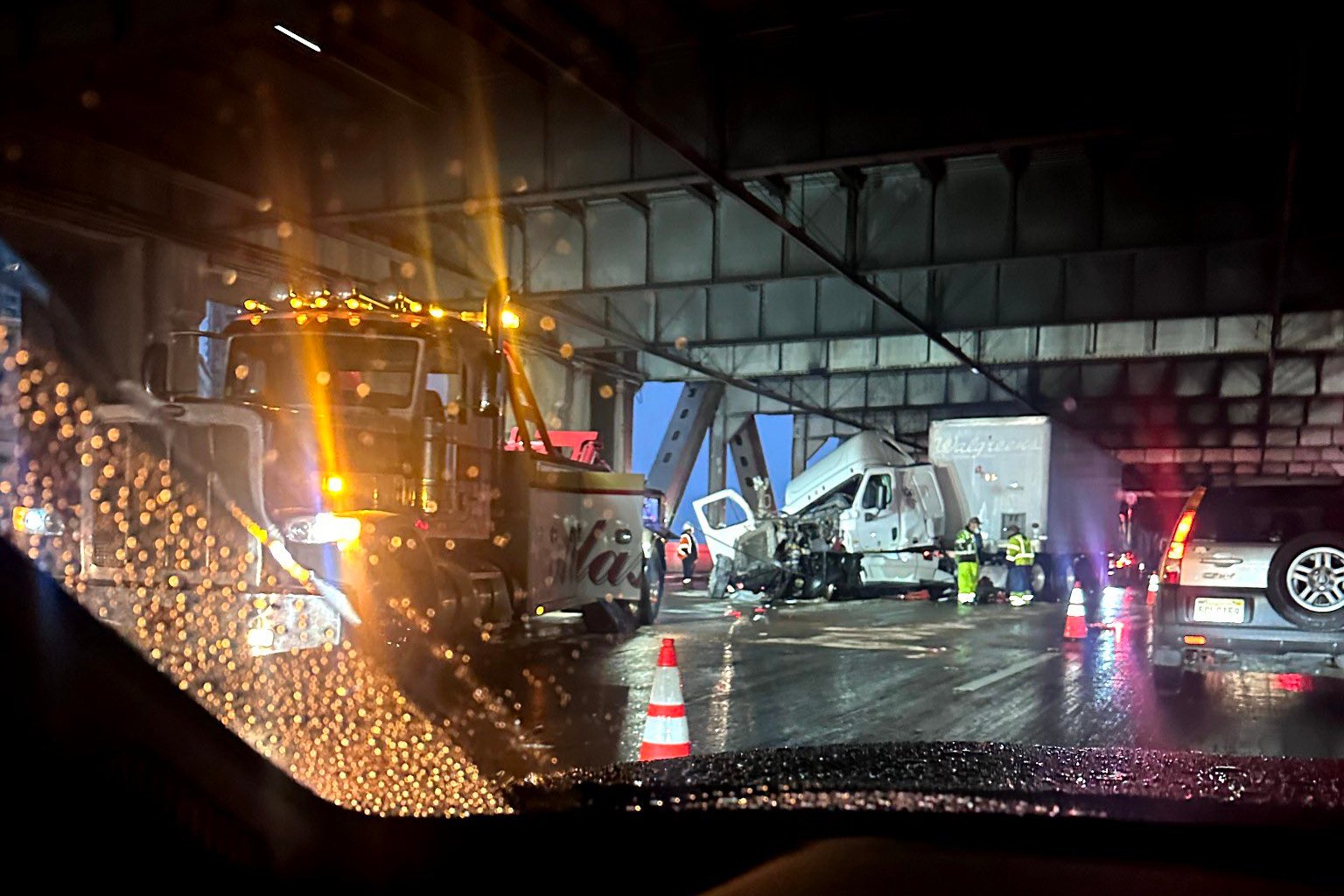 A night scene shows a truck accident under a bridge. Emergency workers are present, and traffic cones are placed around the area. It's raining, and road conditions are wet.