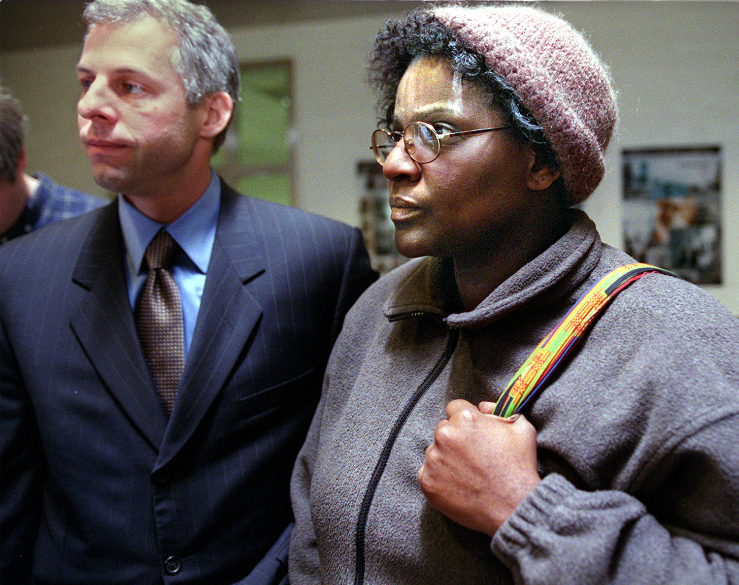 A man in a suit and a woman in glasses and a knit hat stand closely together. The woman holds a strap over her shoulder, and they're in an indoor setting.