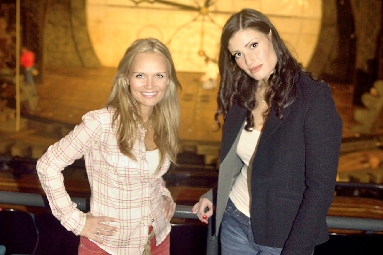 Two women stand smiling in front of a theater stage with a large clock and dragon decoration above. The ambience suggests a set for a theatrical production.