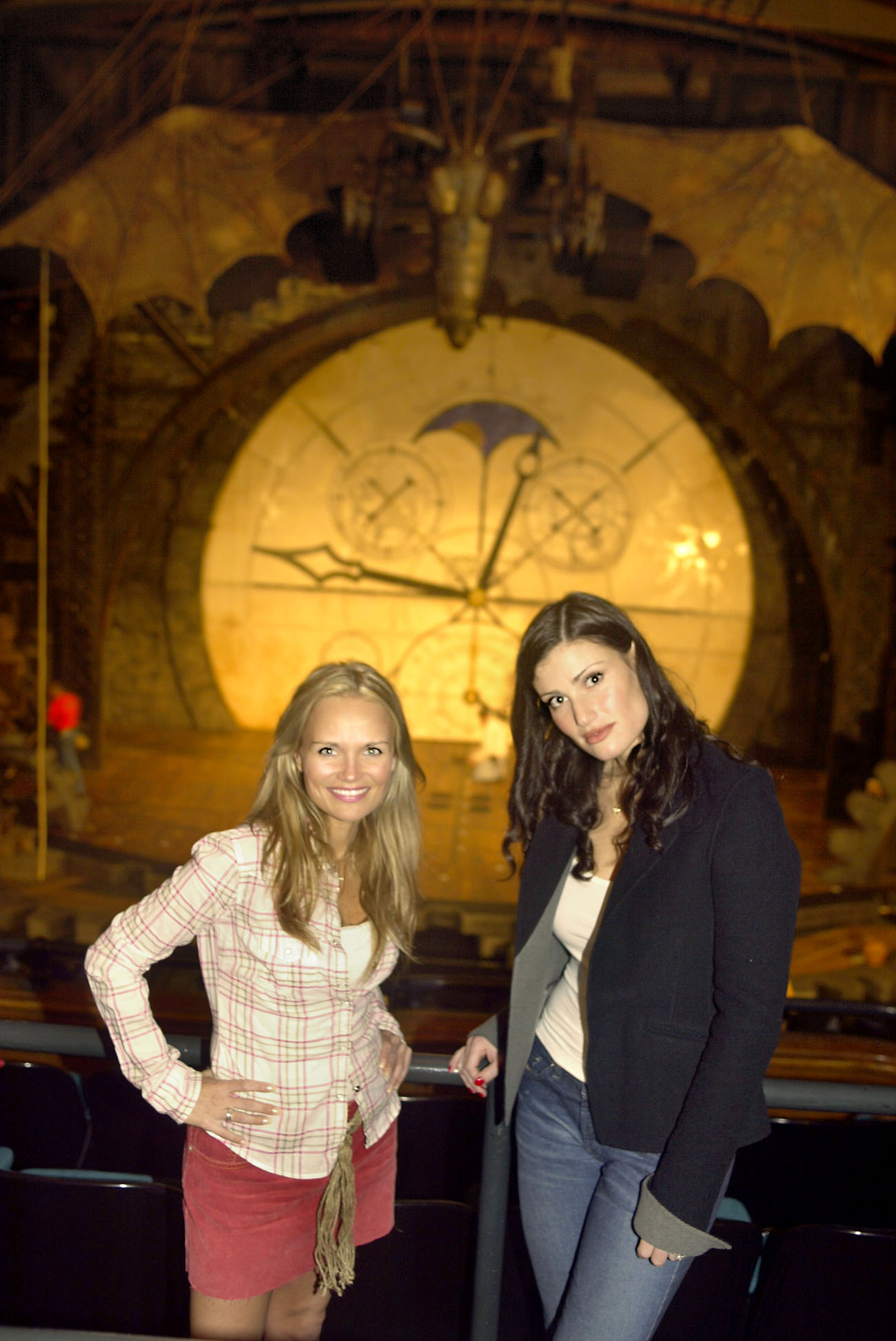 Two women stand smiling in front of a theater stage with a large clock and dragon decoration above. The ambience suggests a set for a theatrical production.