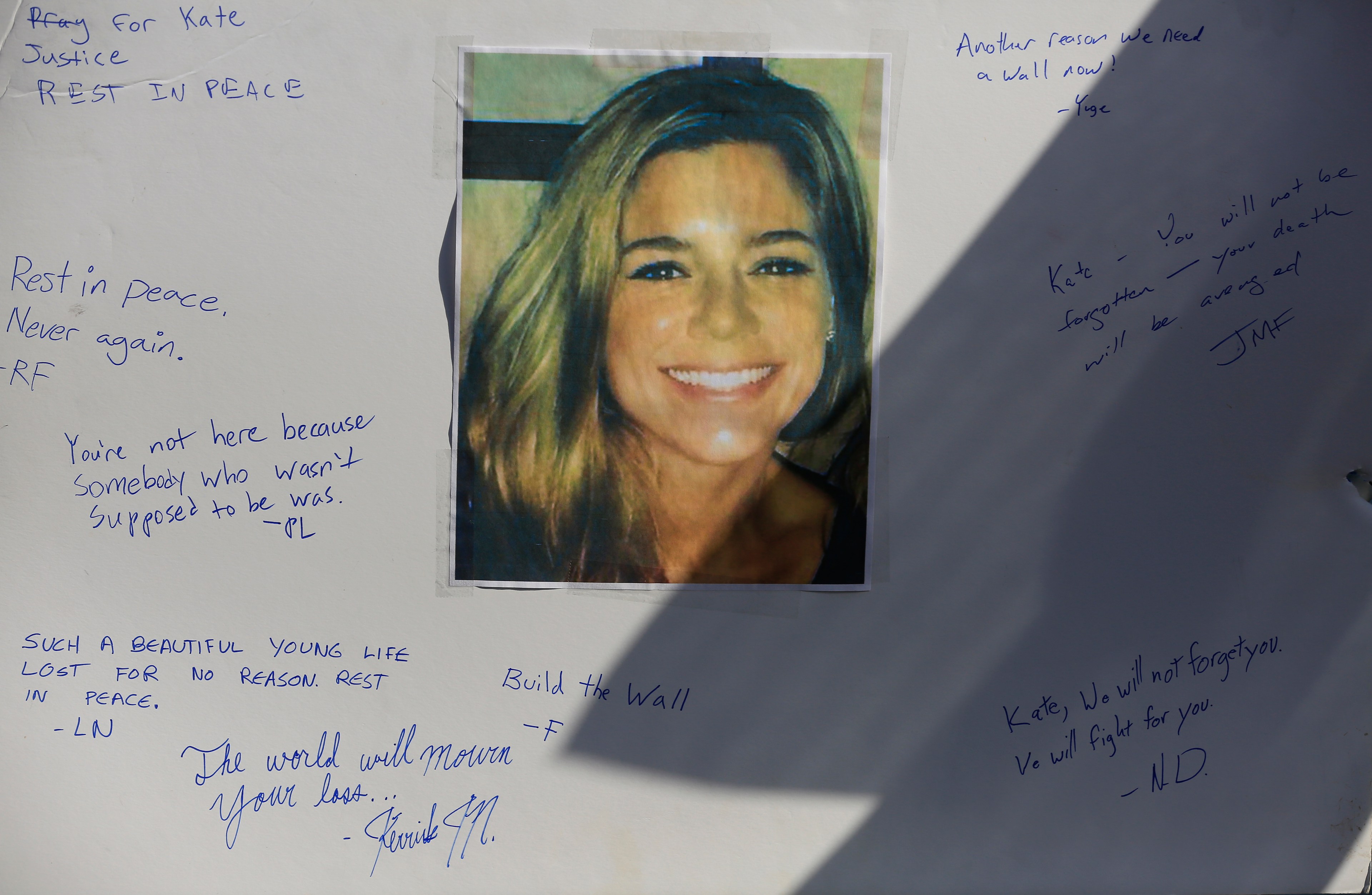 A smiling woman’s photo is taped to a board with handwritten messages surrounding it, expressing condolences and calls for justice with phrases like &quot;Rest in Peace&quot; and &quot;Build the Wall.&quot;