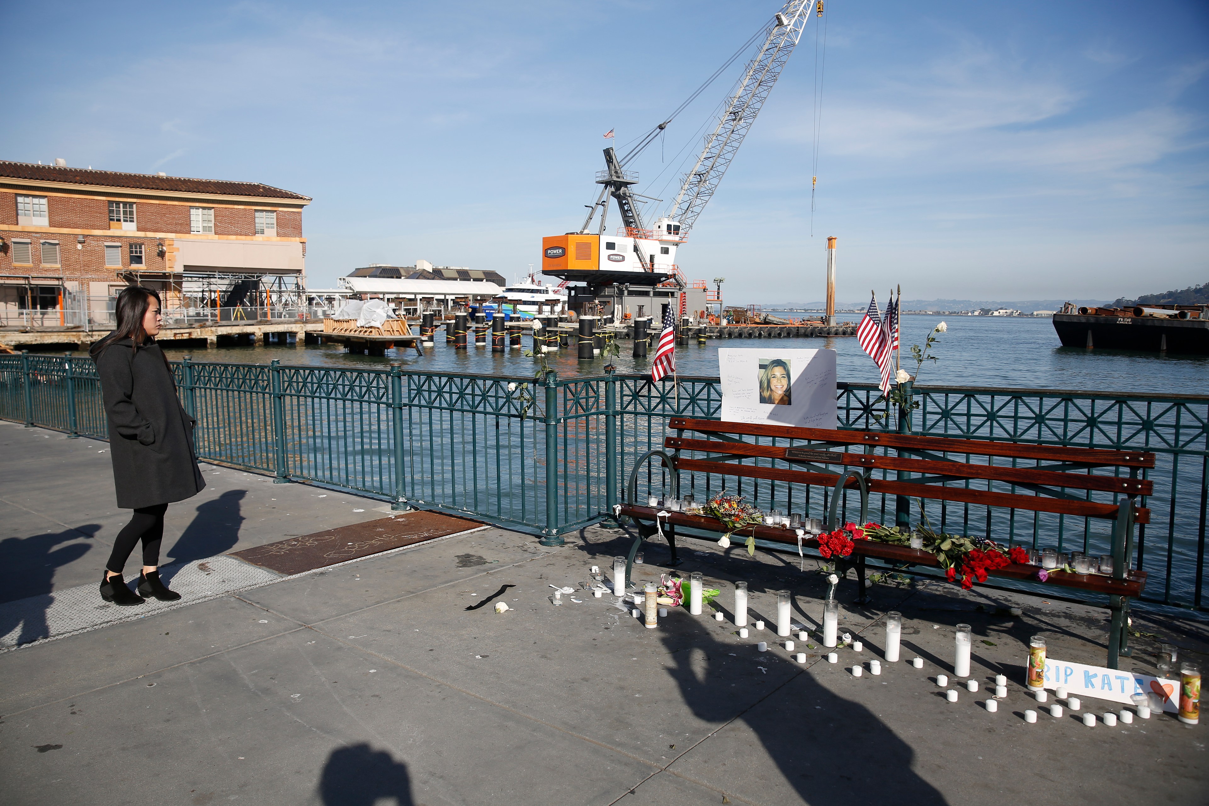 A woman in a dark coat stands near a waterfront memorial, with flowers, candles, and a sign featuring a photo on a bench; a crane and buildings are in the background.