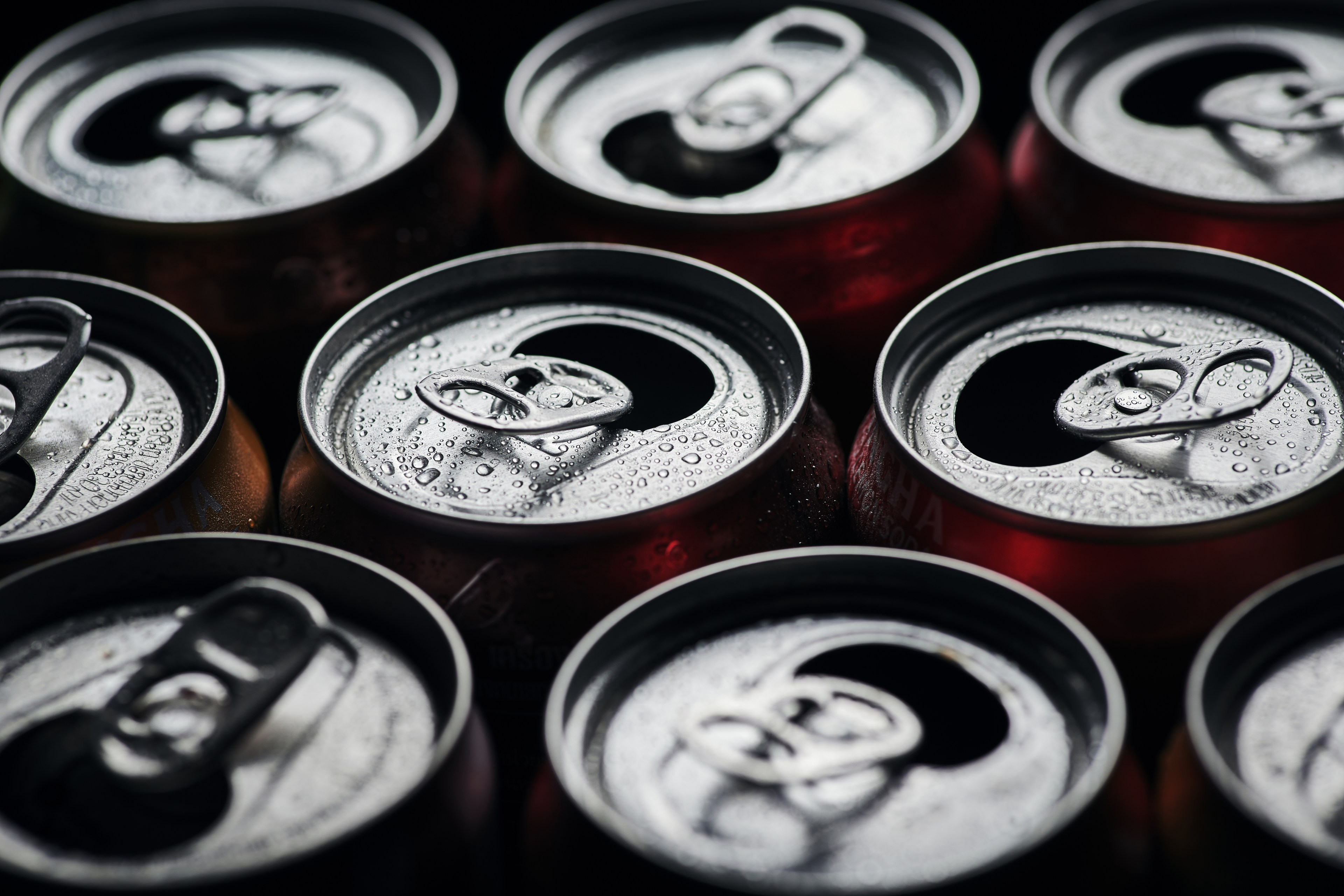 Nine cans with pull tabs are closely positioned, showing droplets of condensation on their surfaces, indicating they're cold and possibly refreshing.