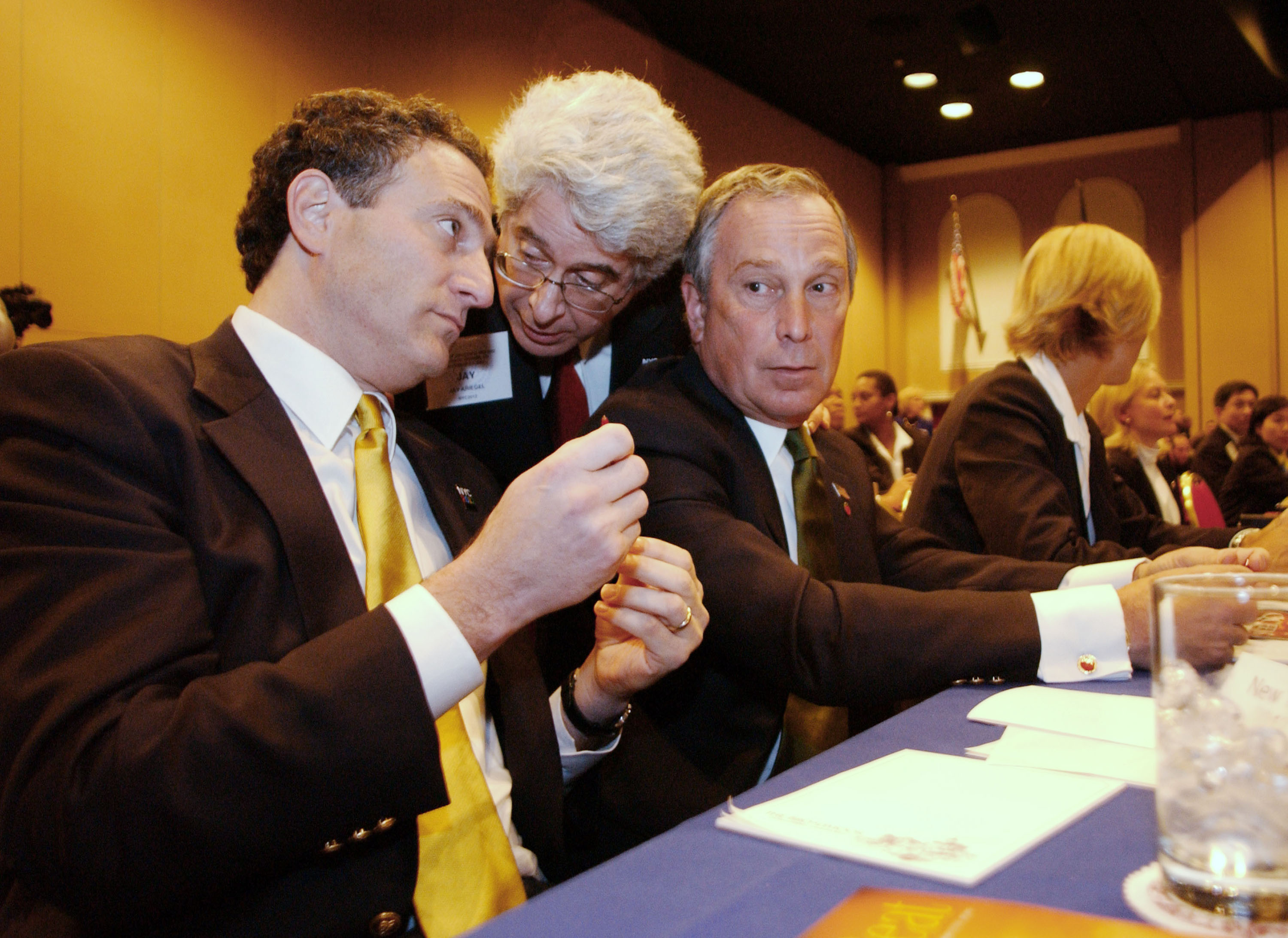 Three men in suits are seated at a table, one whispering to another, while the third listens intently. Papers and a glass of ice water are on the table.