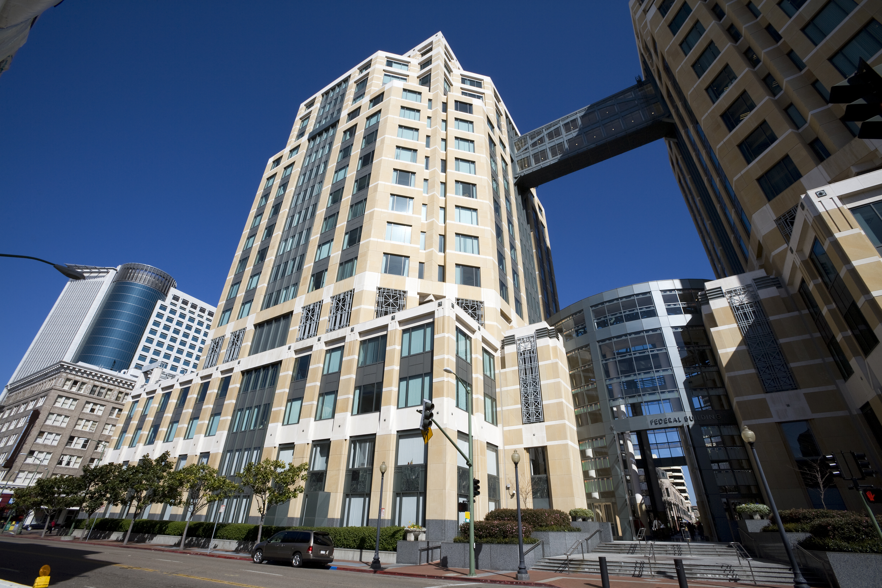 The image shows a tall, modern building with a beige and white exterior. Several rectangular windows line the facade, and a skywalk connects two sections.