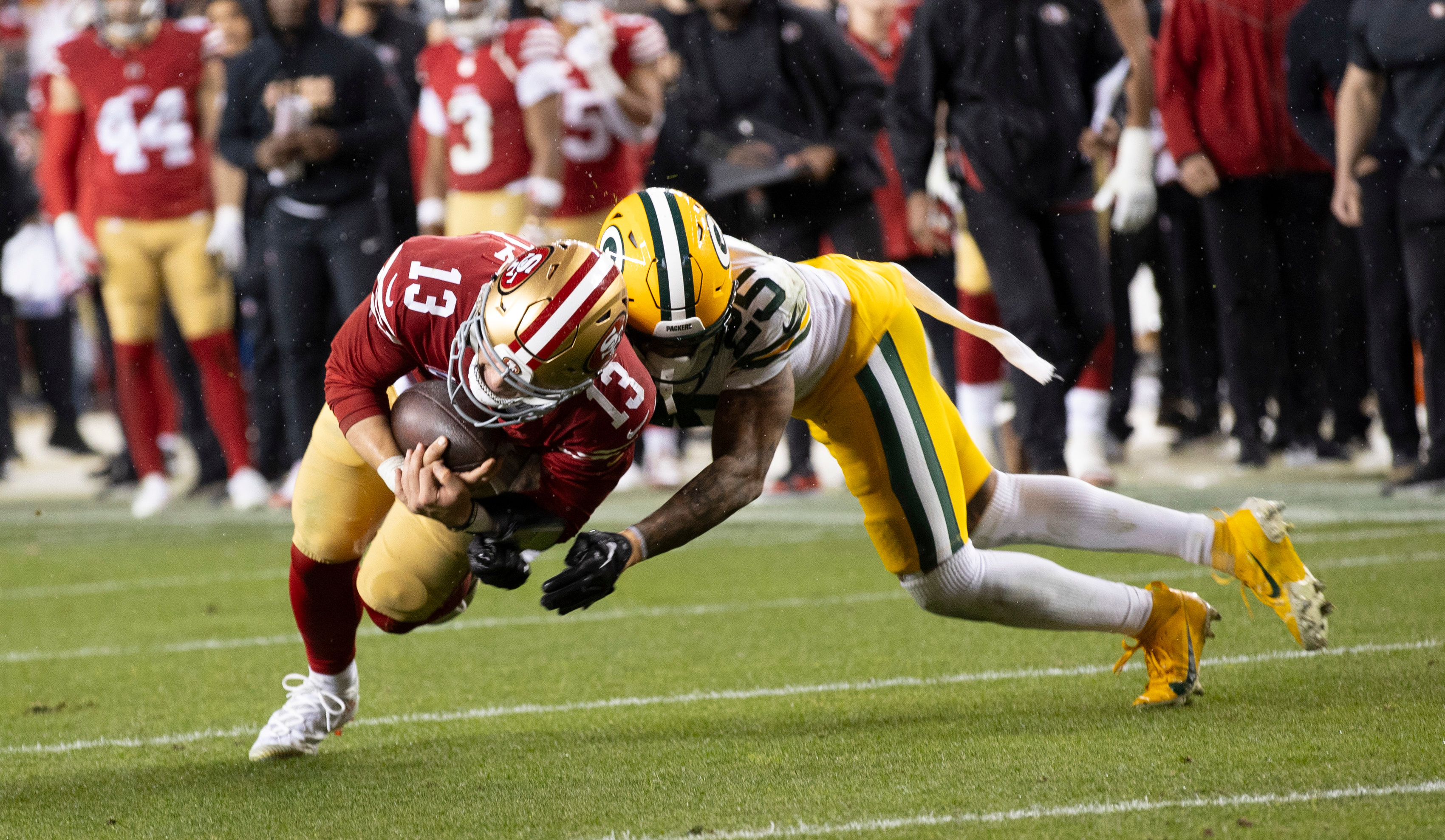 A football player in a red uniform is being tackled by a player in a yellow and green uniform. Both are on a field with blurred spectators in the background.