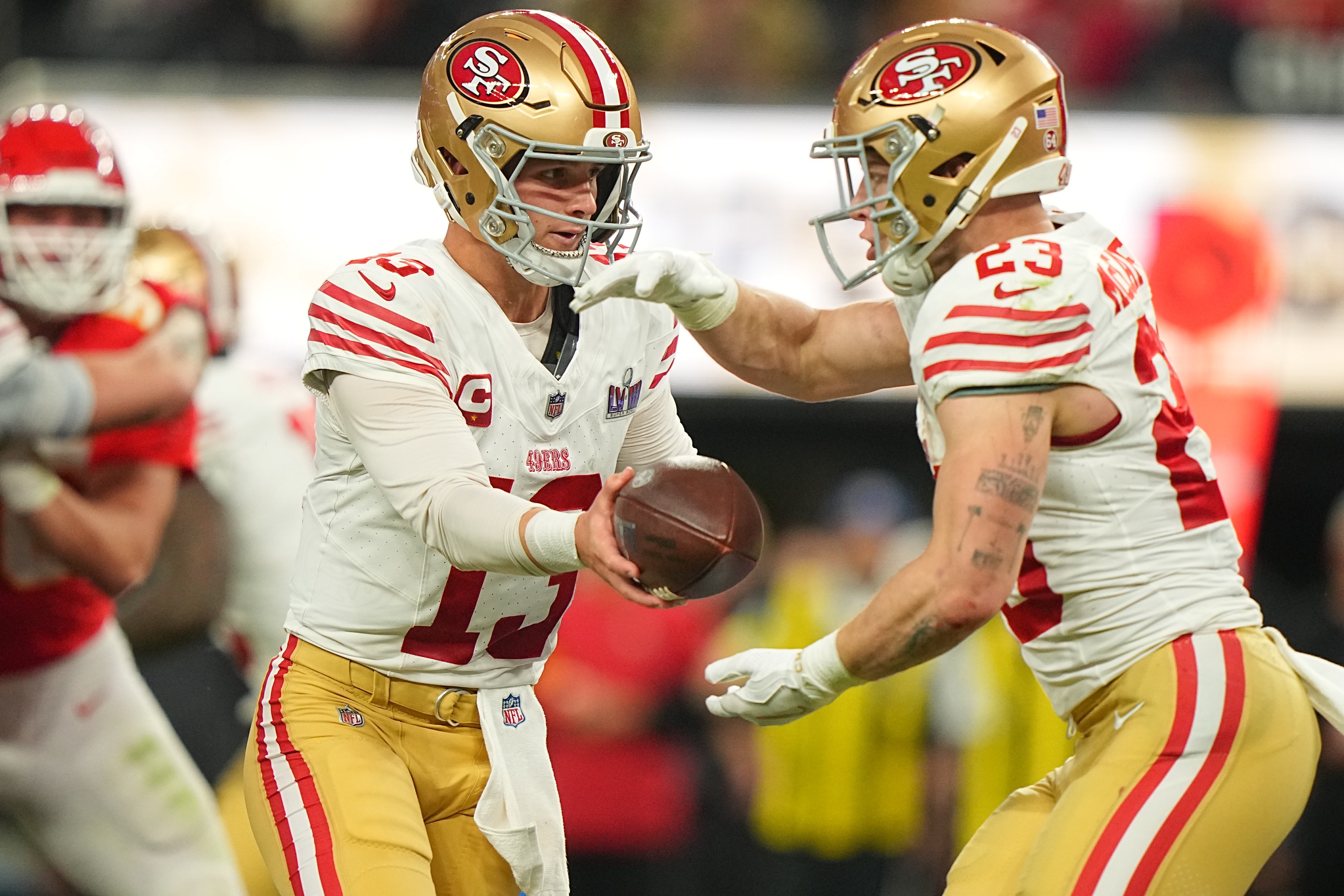 A football player in a gold and red uniform hands the ball to a teammate on the field. They wear helmets and are engaged in an intense game.