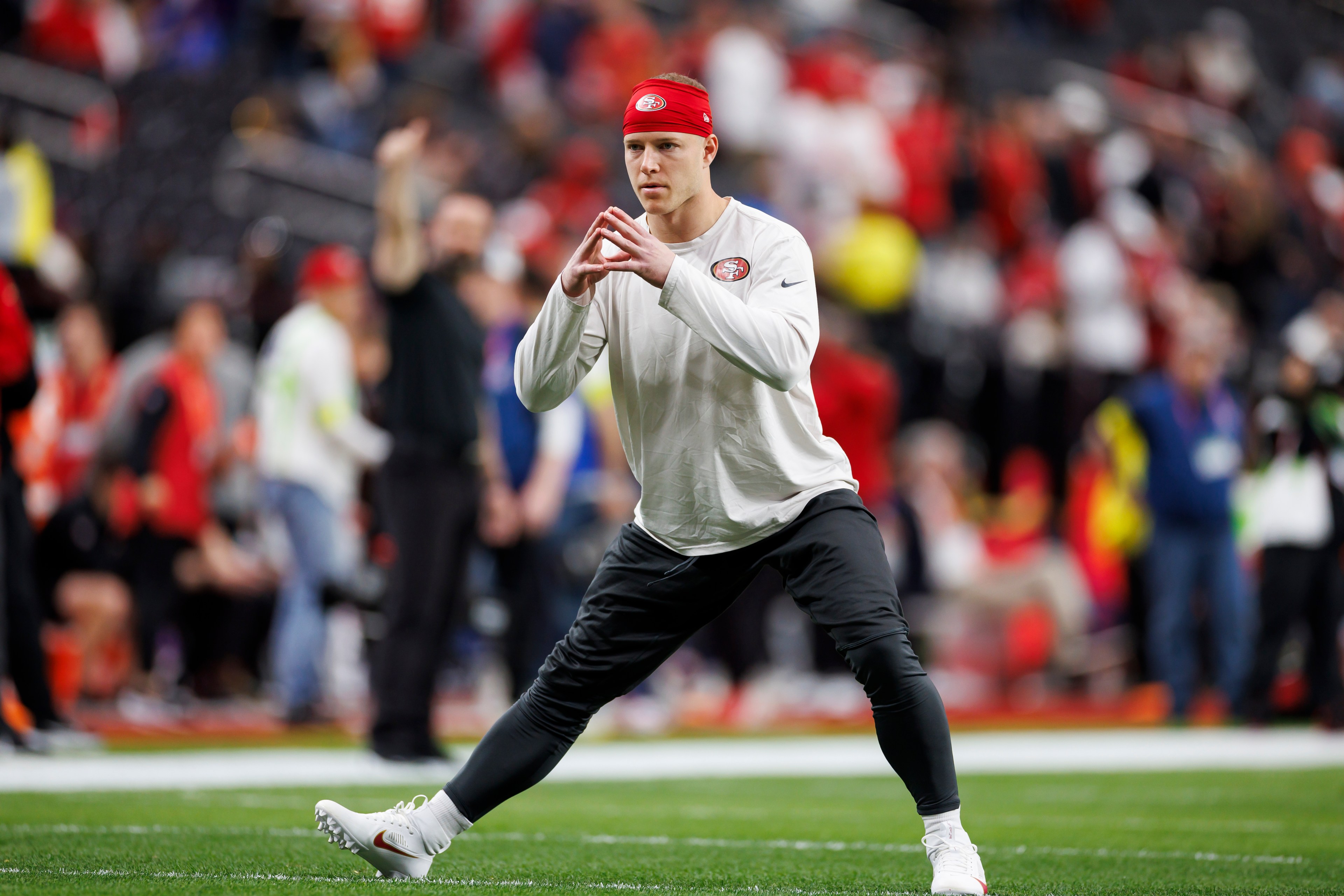 A man in a white long-sleeve shirt and red headband stretches on a football field. He's focusing, with hands clasped, while people in the background watch.