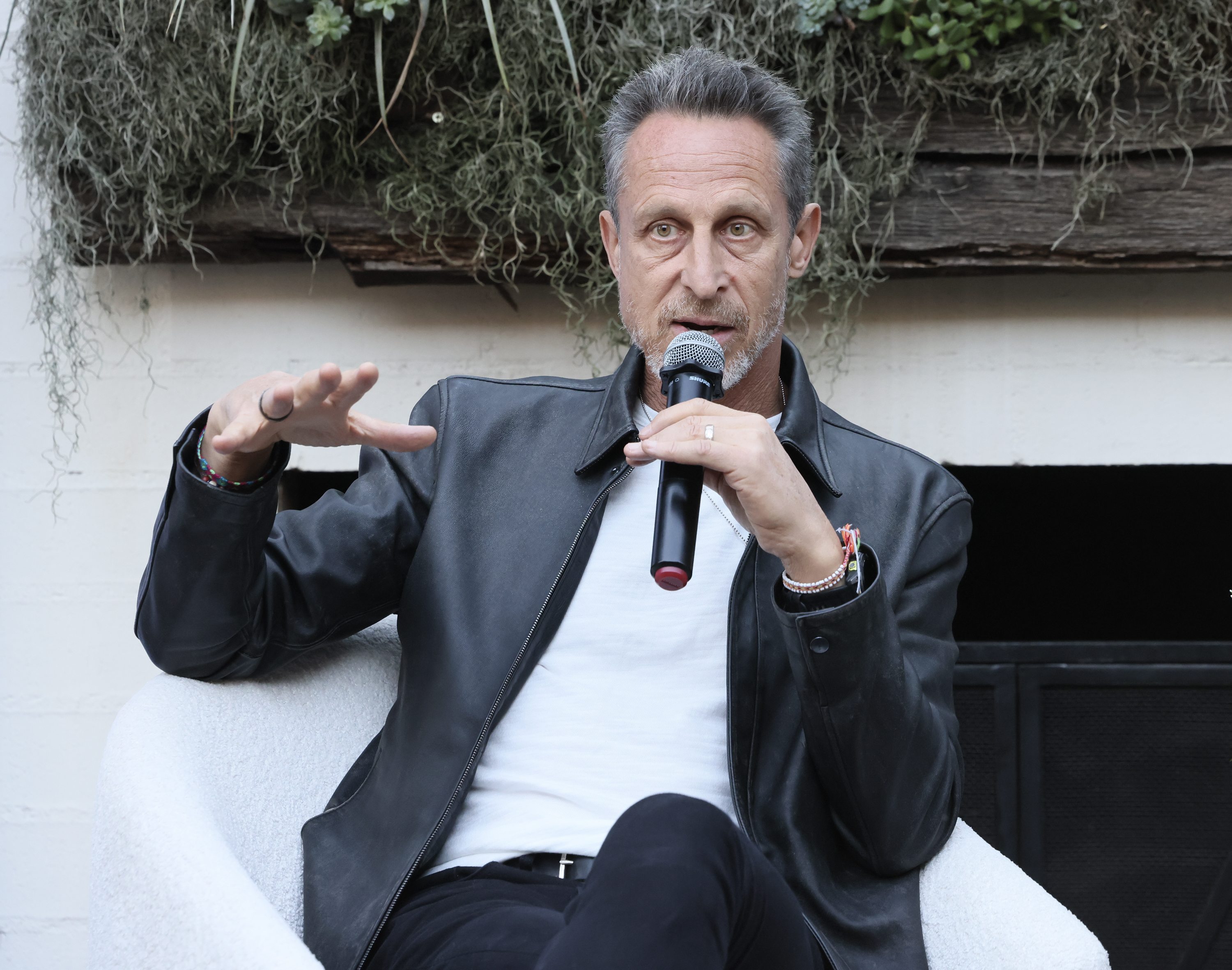 A man in a leather jacket speaks into a microphone while seated in a white chair. He gestures with one hand, and there is greenery hanging on the wall behind him.