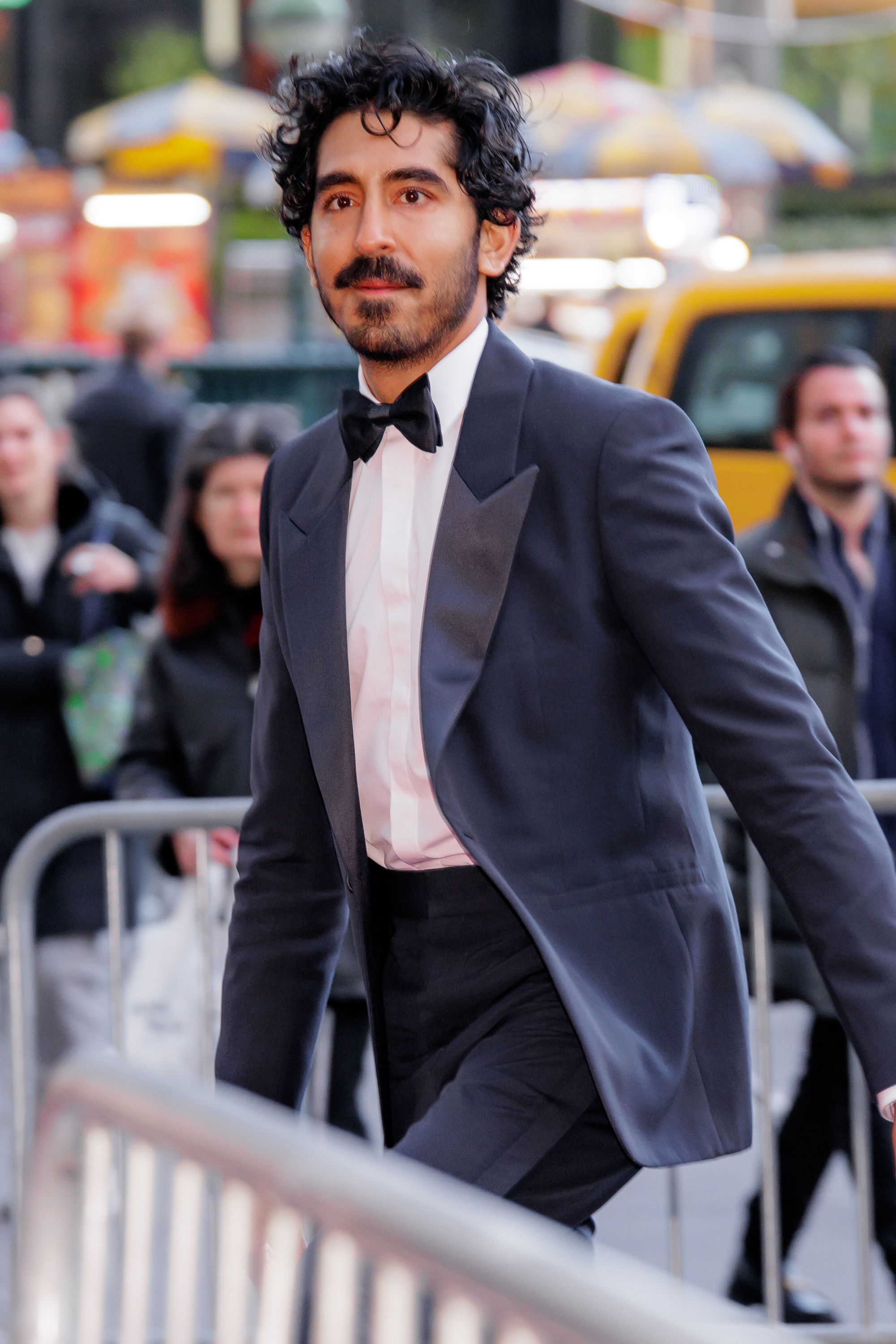 A man with curly hair and a mustache walks in a tuxedo with a bow tie. People and a yellow taxi are visible in the blurred background.