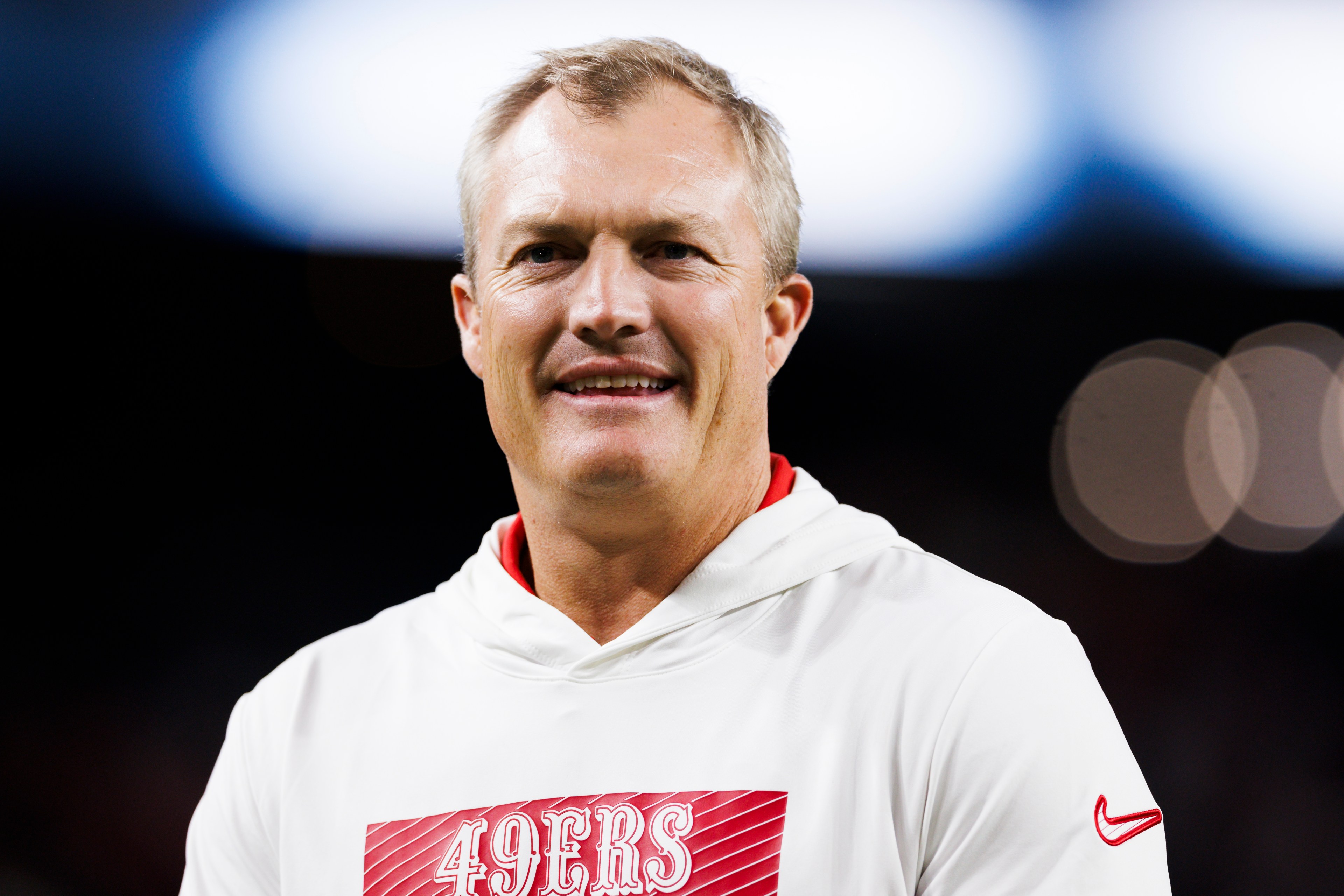 A smiling man is wearing a white hoodie featuring a red panel with "49ERS" on it. The background is blurred, with bright, out-of-focus lights.