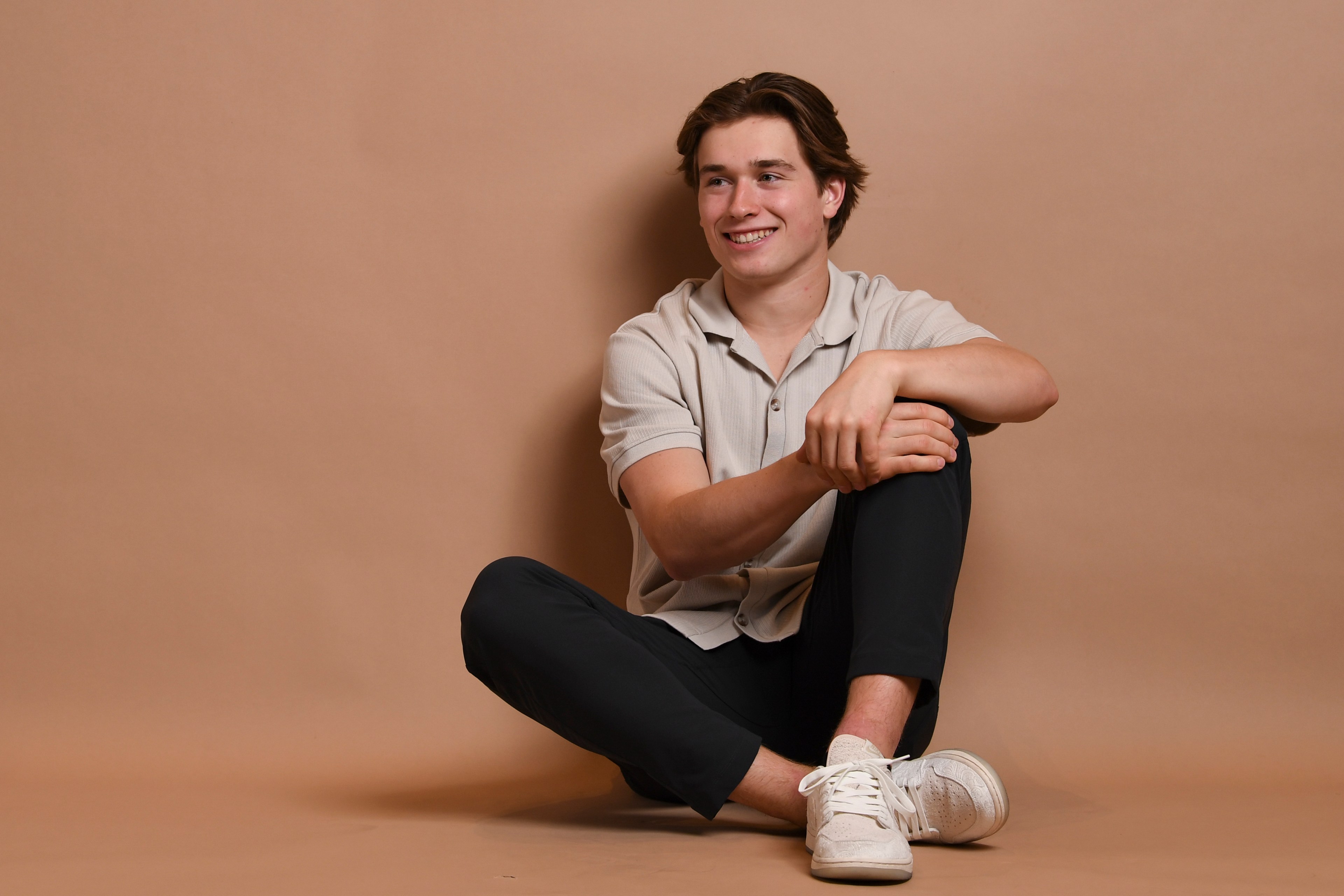 A person is sitting cross-legged on the floor against a beige background, smiling. They wear a light shirt, dark pants, and white sneakers, appearing relaxed and casual.