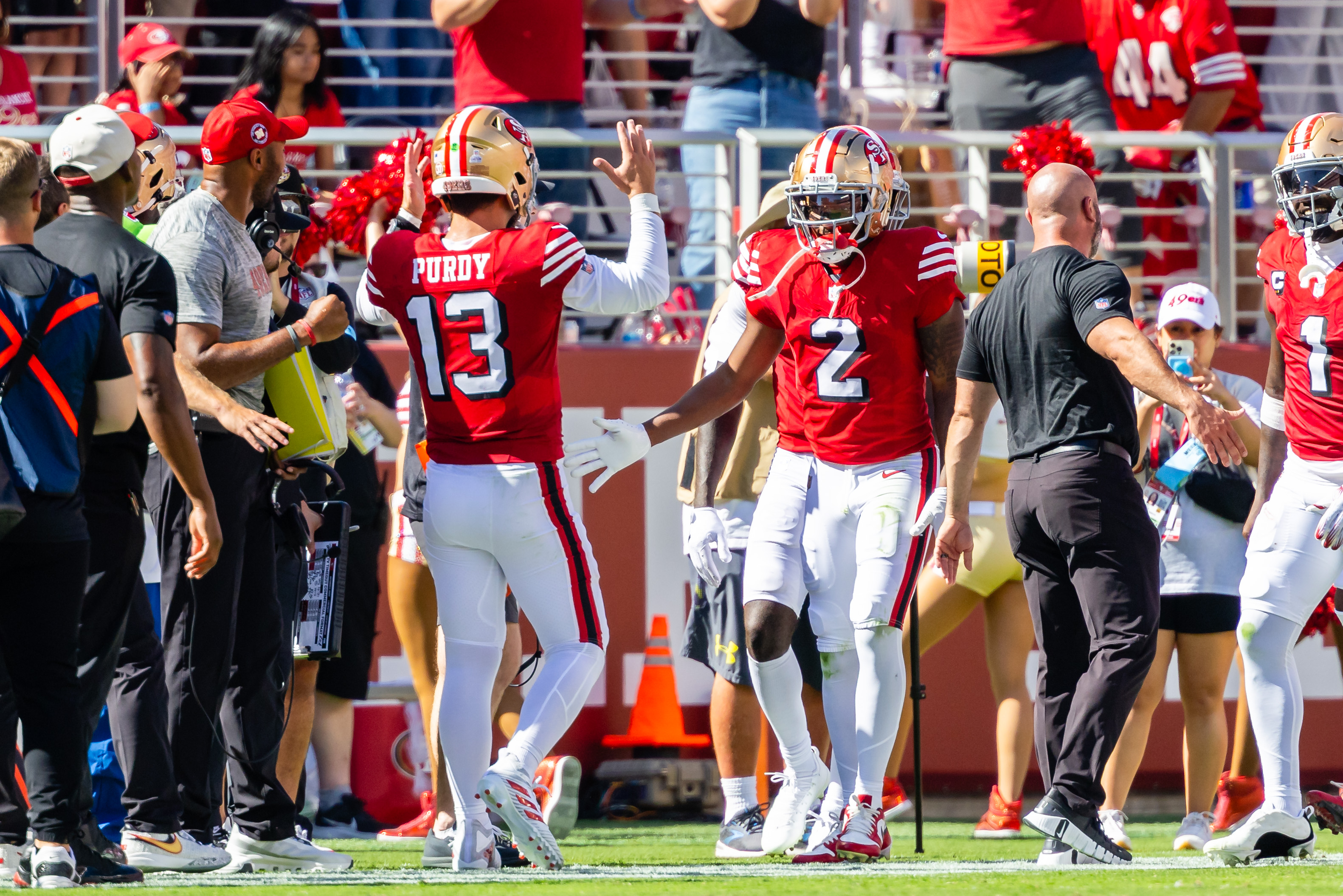 A football player in a red jersey with the number 13 high-fives a teammate wearing number 2. They're surrounded by coaches and staff on the sidelines.