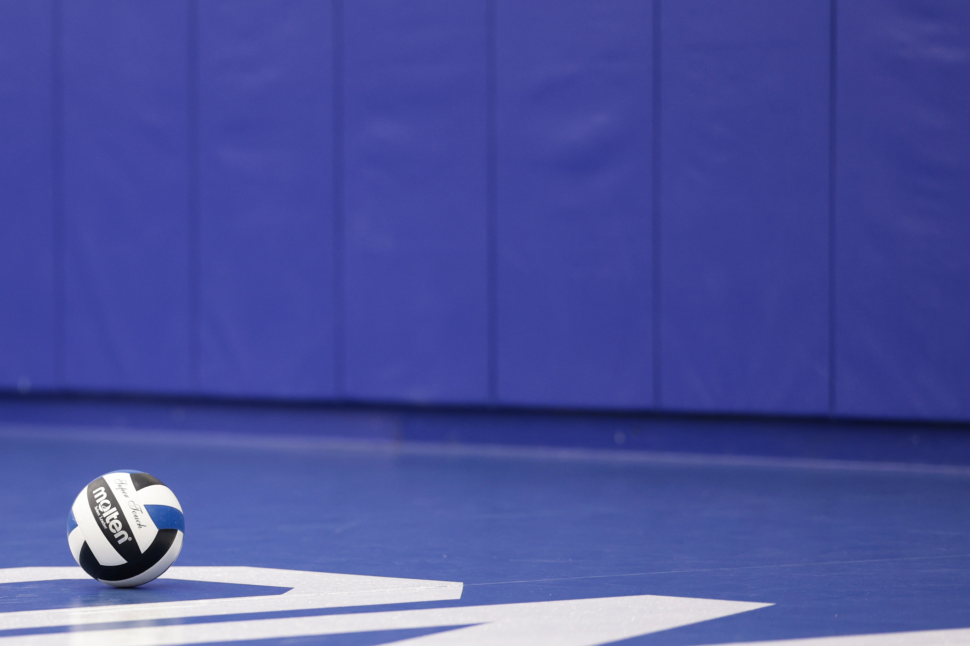 A volleyball rests on a blue gym floor with a padded blue wall in the background. The ball is black, white, and blue with the brand name visible.