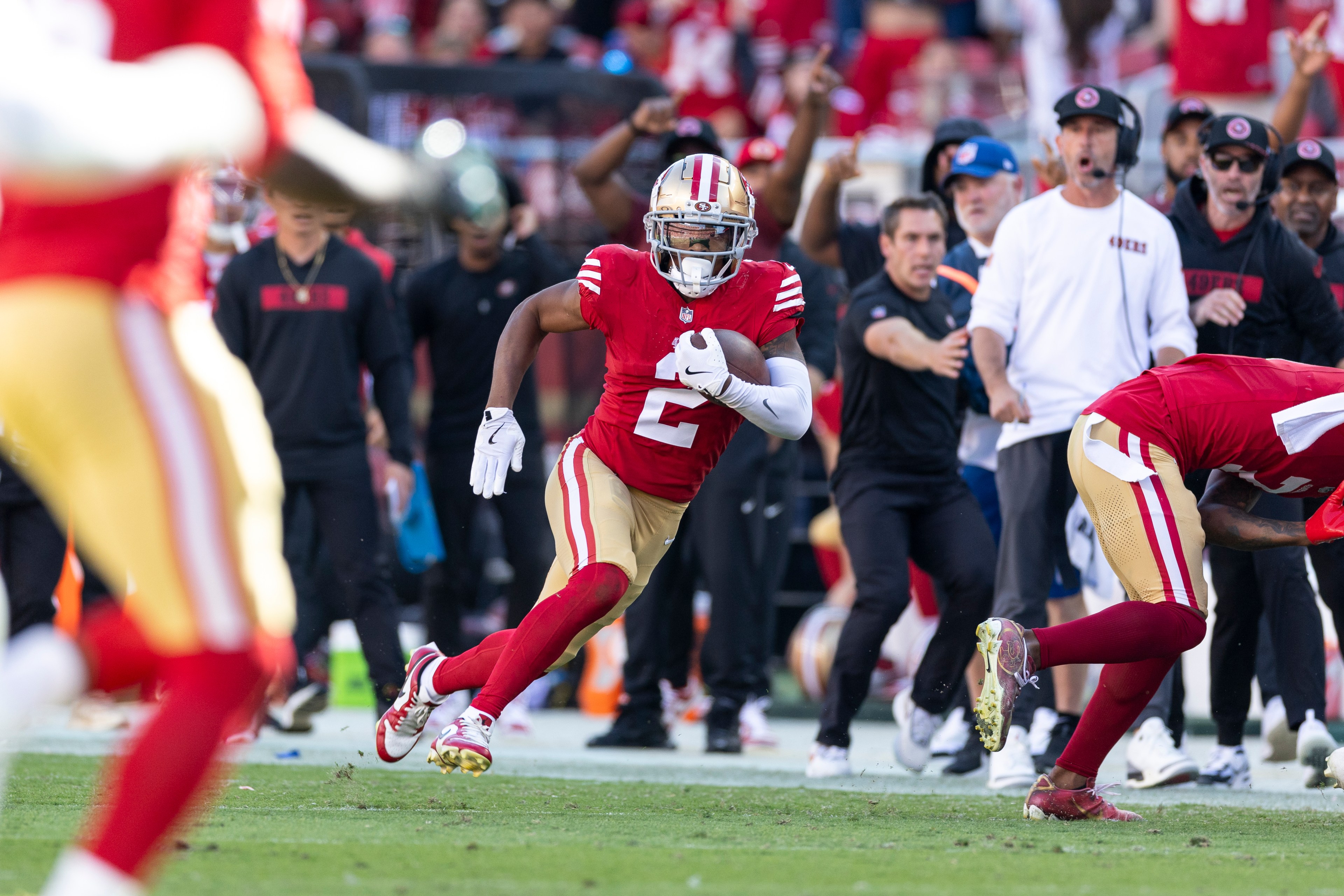 A football player in a red jersey runs with the ball on a field, closely followed by teammates. Coaches and staff on the sidelines react in the background.