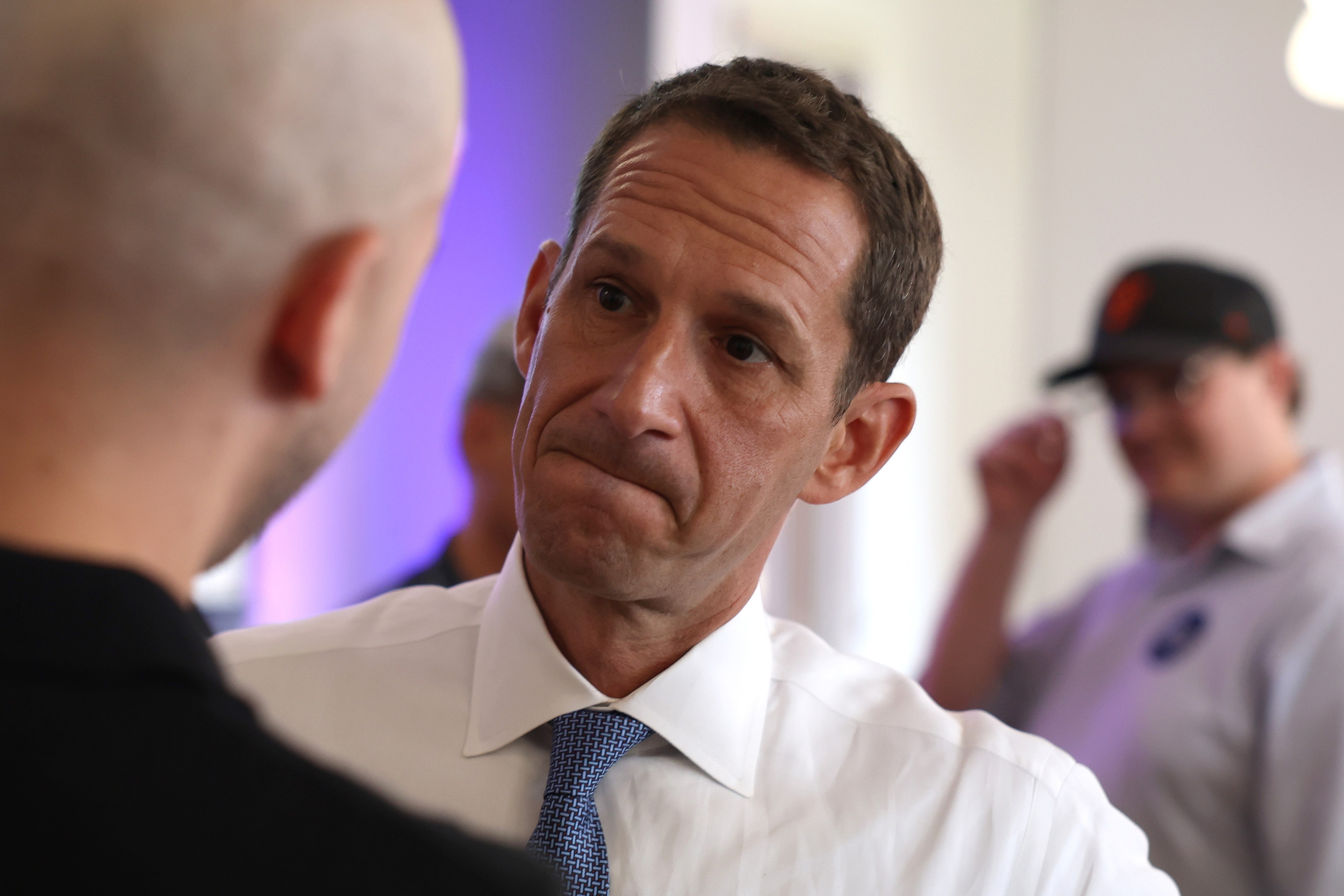 A man in a white shirt and blue tie looks attentively at another person with a shaved head. In the blurred background, another person wears a cap and glasses.