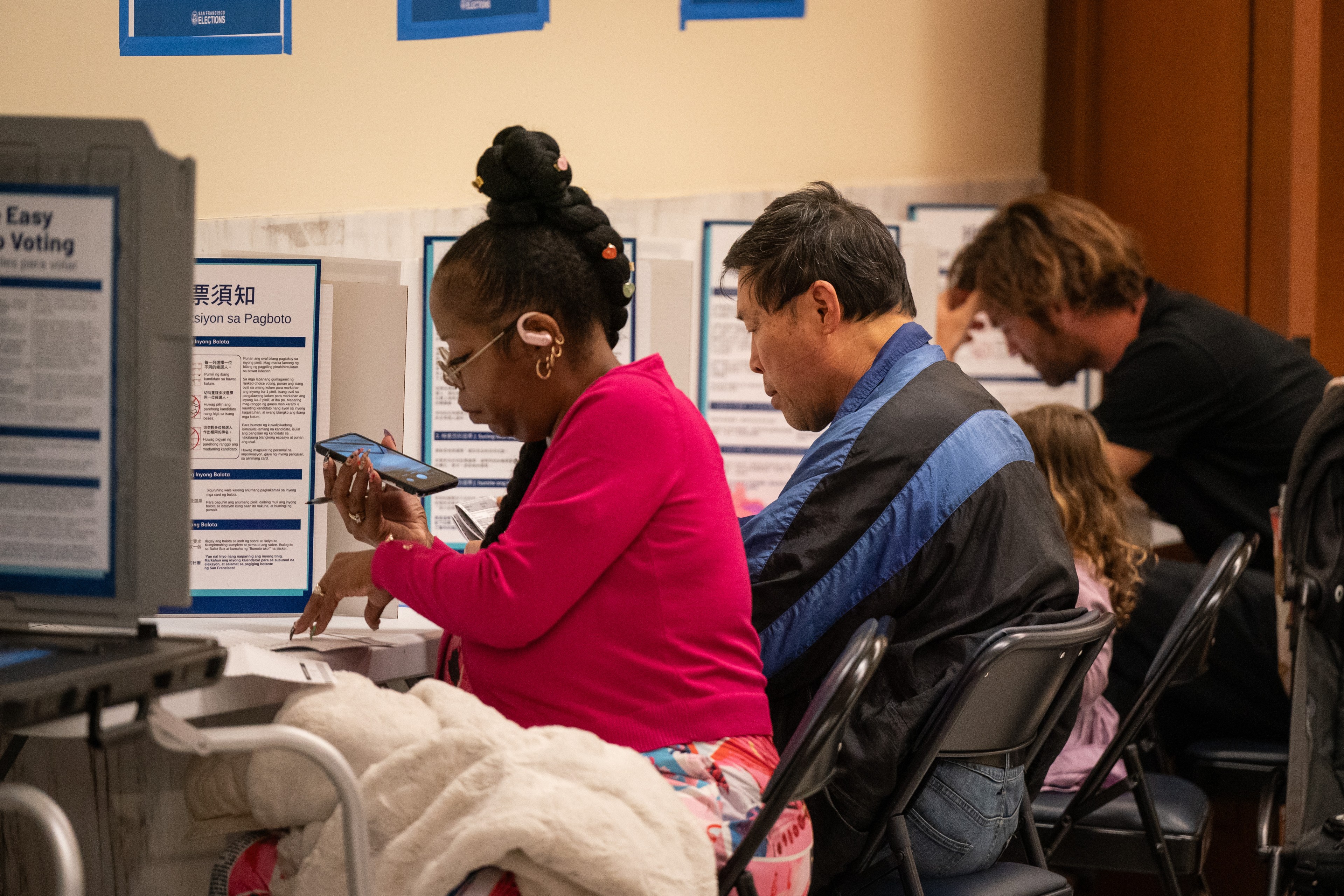 People are sitting in a row at voting booths, focused on filling out ballots. One person holds a phone and wears headphones, indicating accessibility needs.