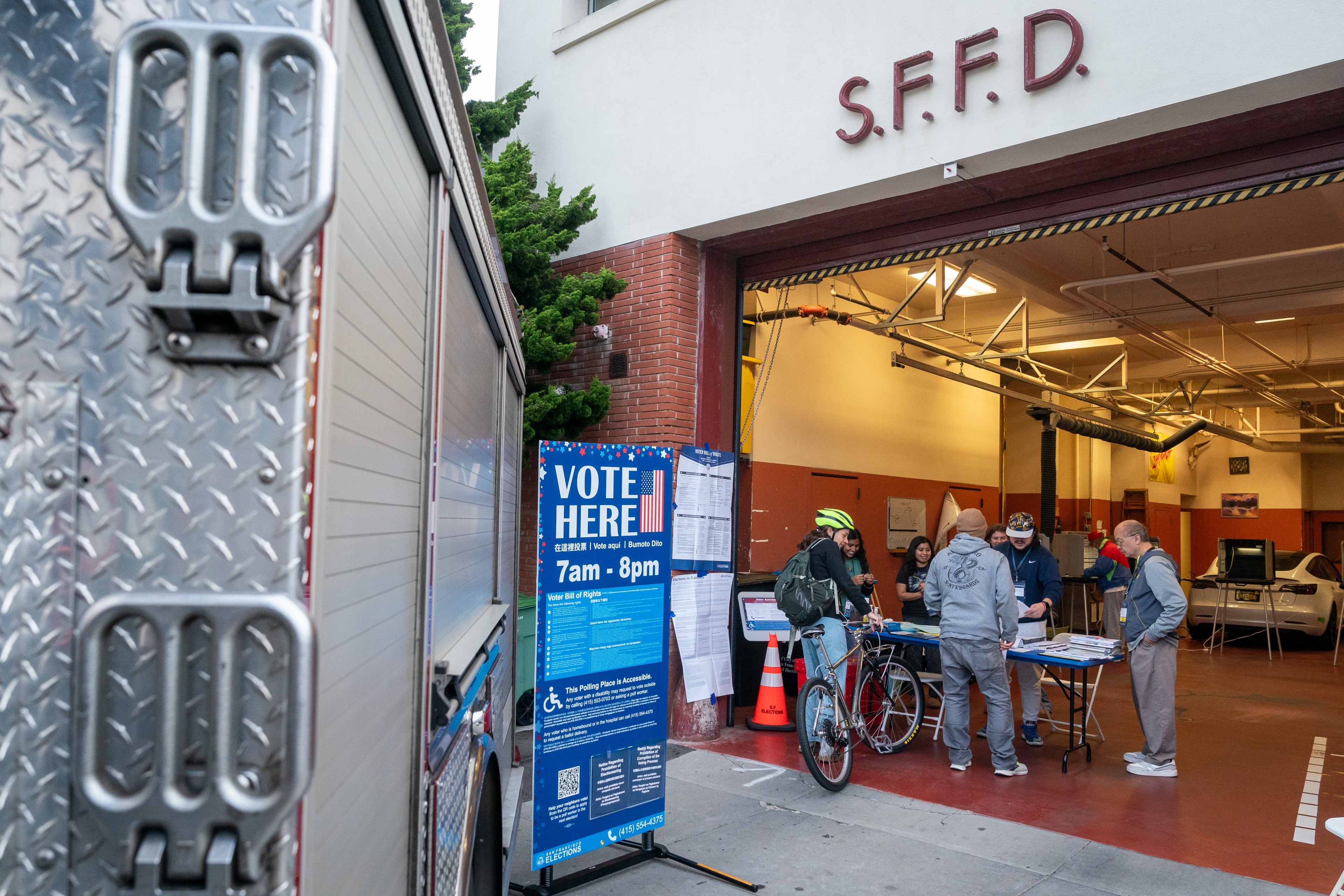 People are gathered outside an S.F.F.D. building, voting at a table with signs and a bicycle nearby. A &quot;VOTE HERE&quot; sign indicates polling hours.