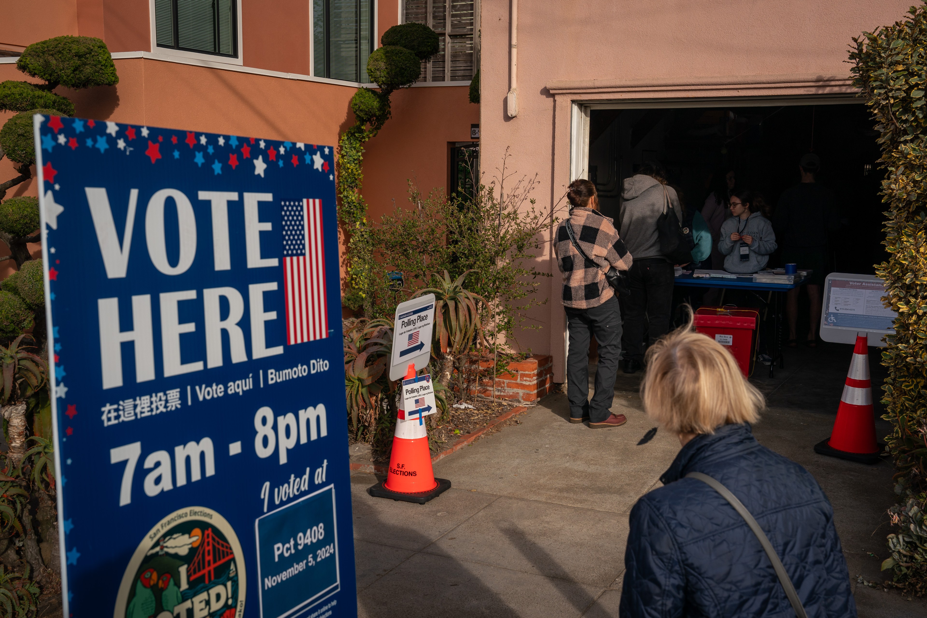 People line up at a polling place in a residential area. A &quot;Vote Here&quot; sign in multiple languages and election cones are visible.