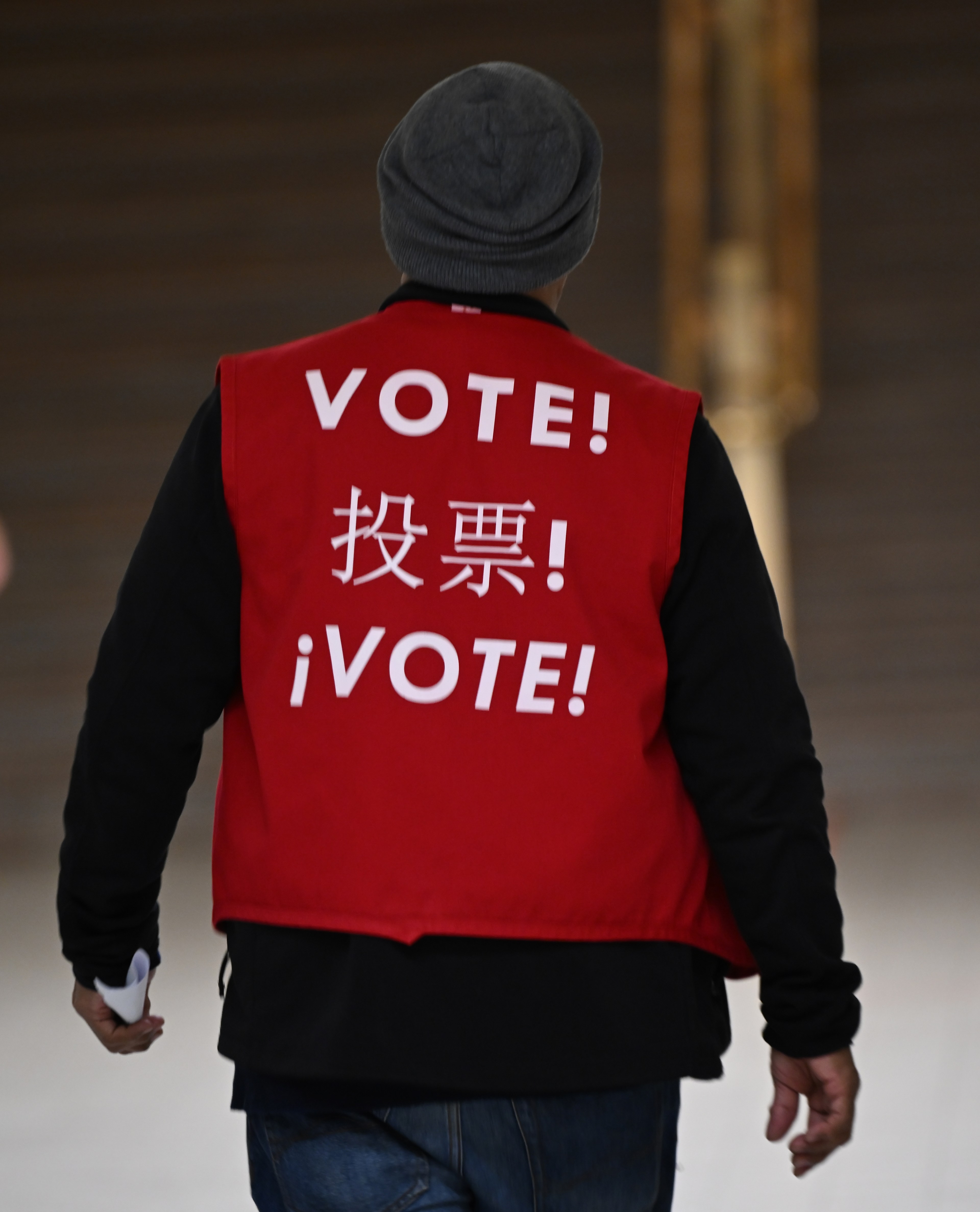 A person wearing a red vest with the word &quot;VOTE!&quot; in English, Chinese, and Spanish. They are walking away, holding some papers, and wearing a gray beanie.