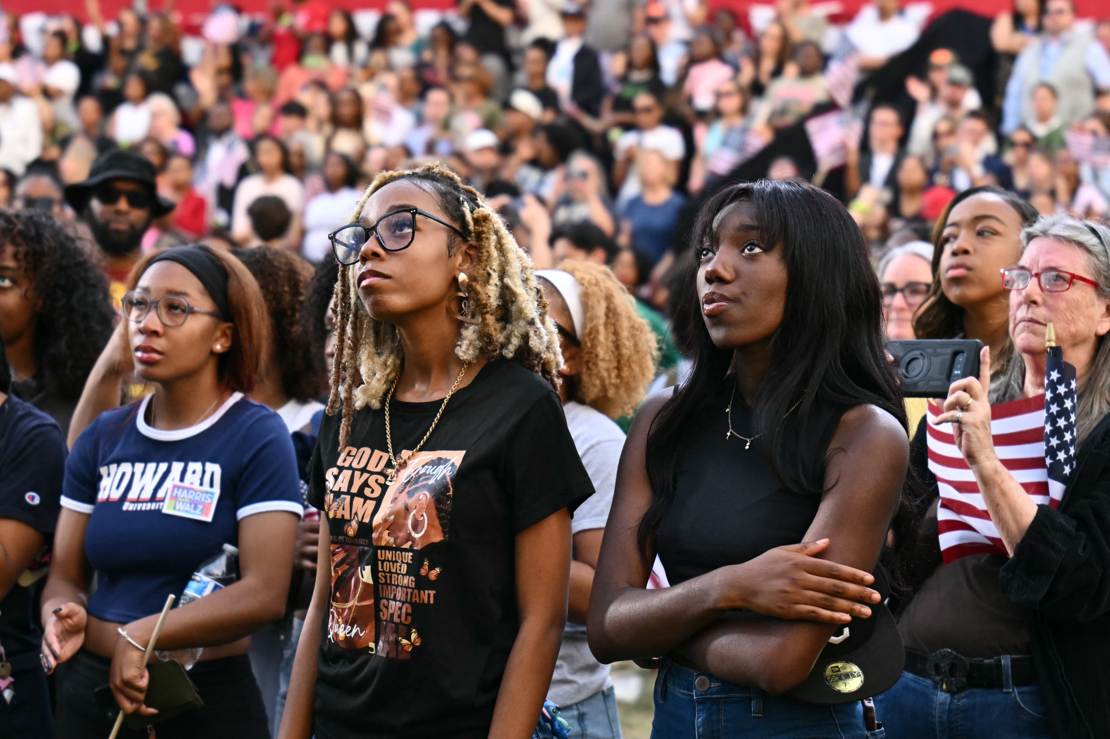 A diverse crowd attentively watches an event, with people wearing various shirts, holding small American flags, and capturing moments on their phones.