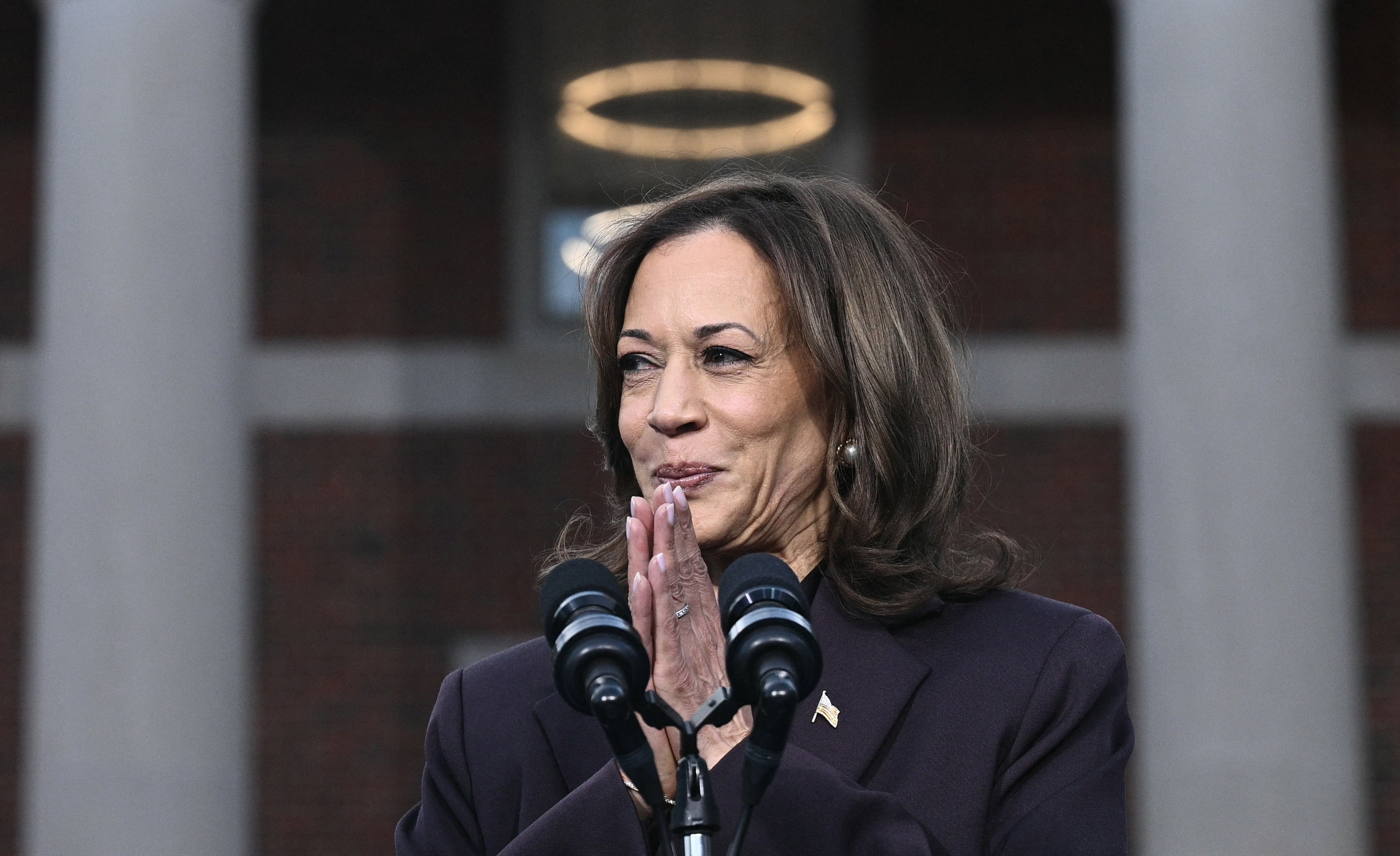 A woman is speaking at a podium with microphones. She appears thoughtful, with hands clasped in front of her face. A circular light is visible in the background.