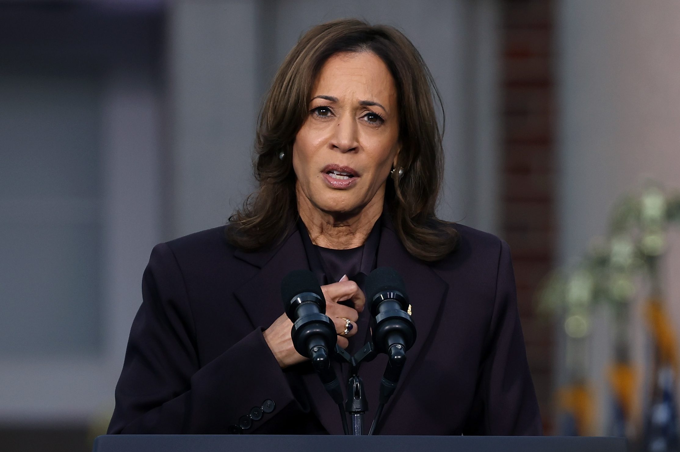 A woman in a dark suit speaks at a podium with "Vice President of the United States" seal. U.S. flags are blurred in the background, and she's speaking into microphones.