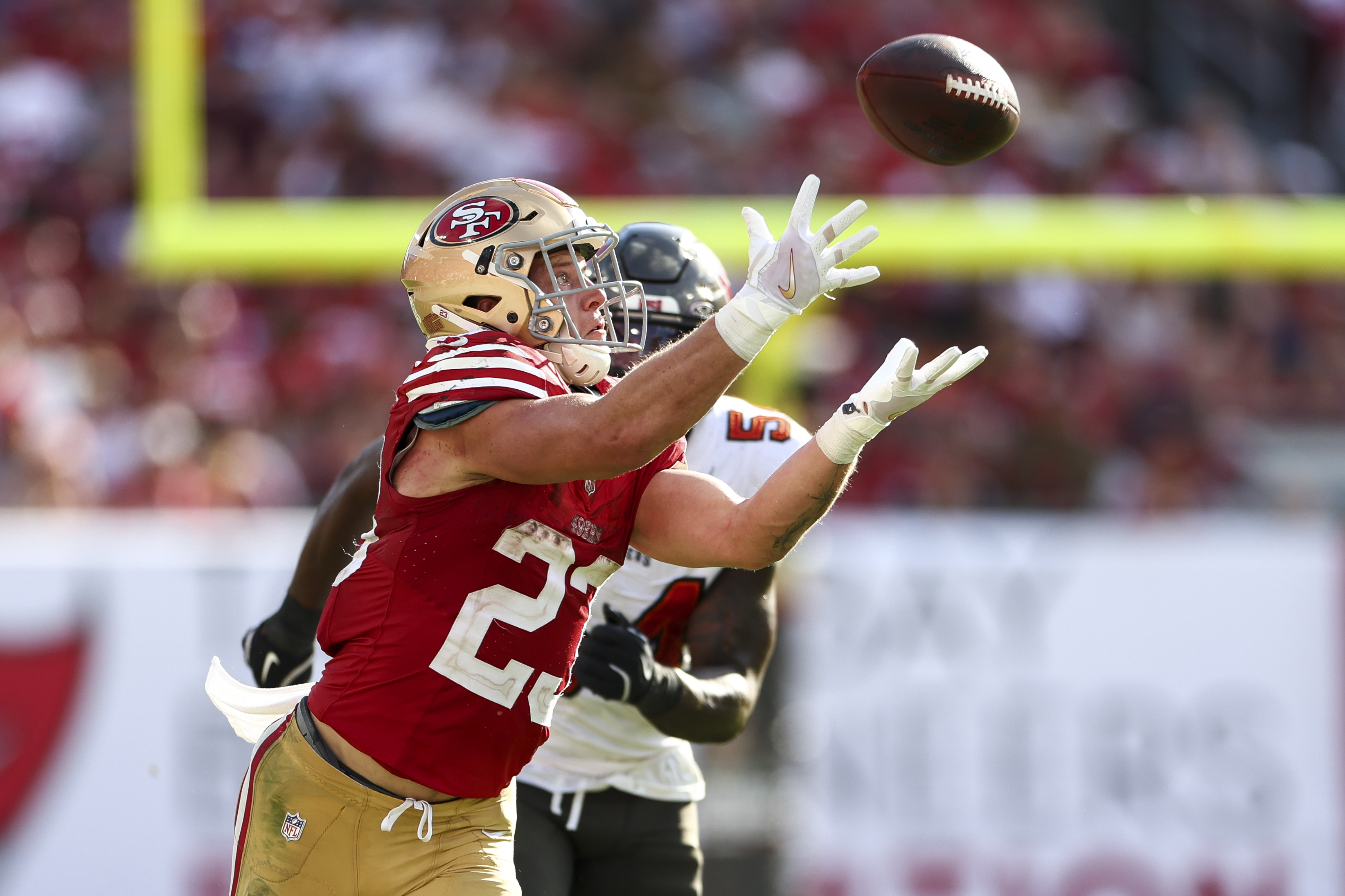 A football player in a red and gold uniform reaches to catch a ball mid-air, with an opposing player closely behind him on the field.