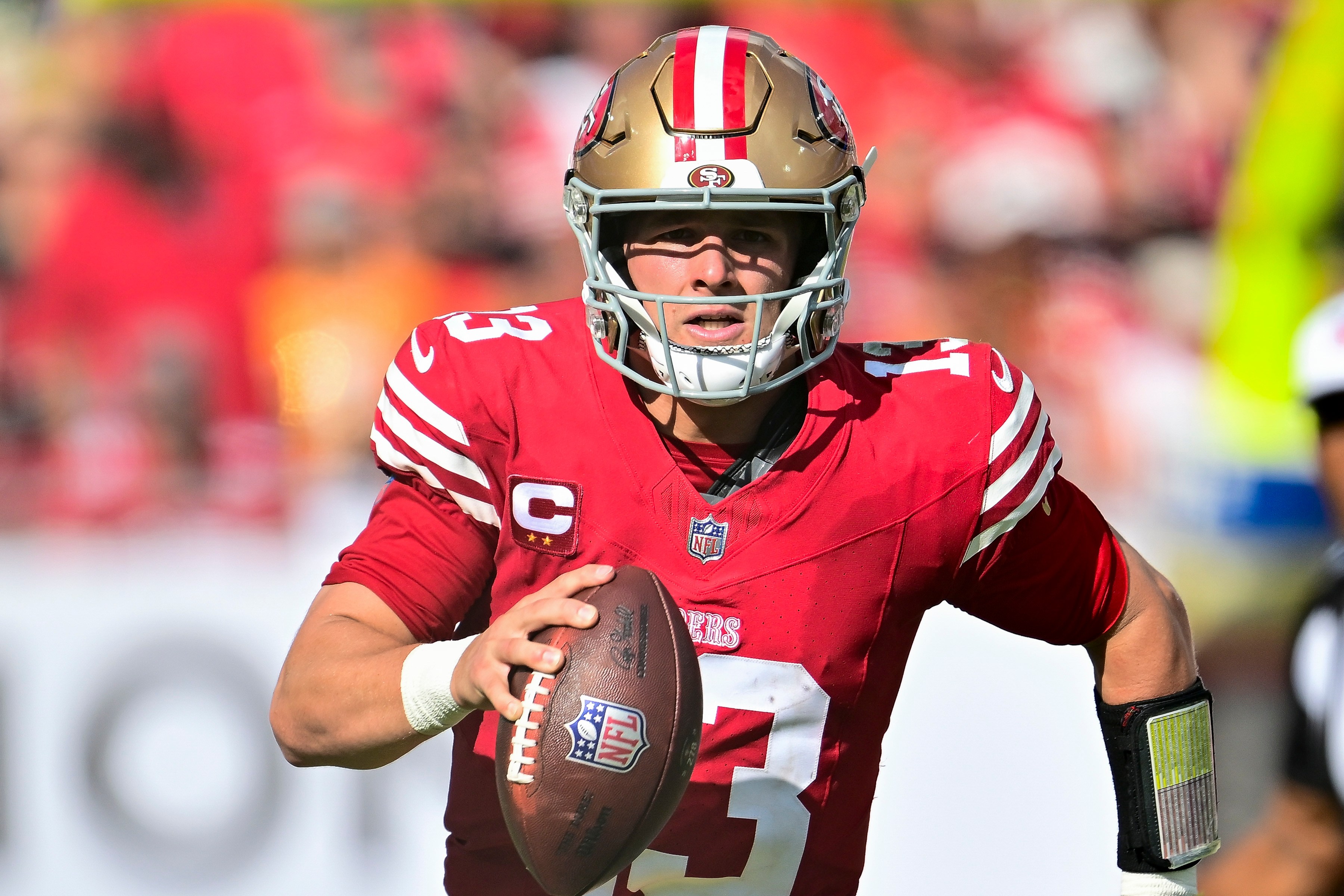 A football player in red and gold uniform runs on the field, holding the ball. He's wearing a helmet, and sunlight shines on him, creating a dynamic action scene.