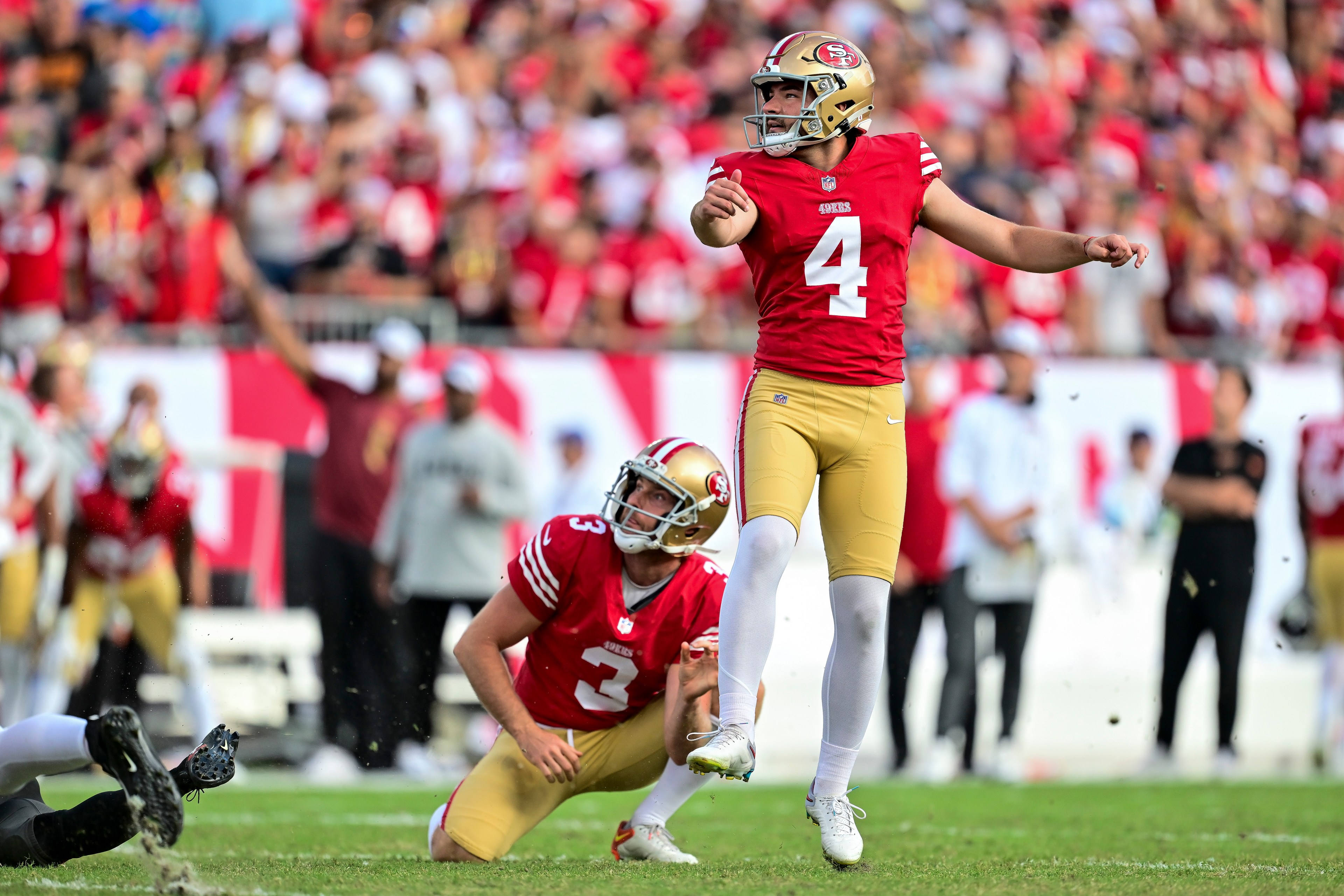 A football player in a red and gold uniform, number 4, is kicking a ball with a teammate, number 3, holding it on a field surrounded by a cheering crowd.