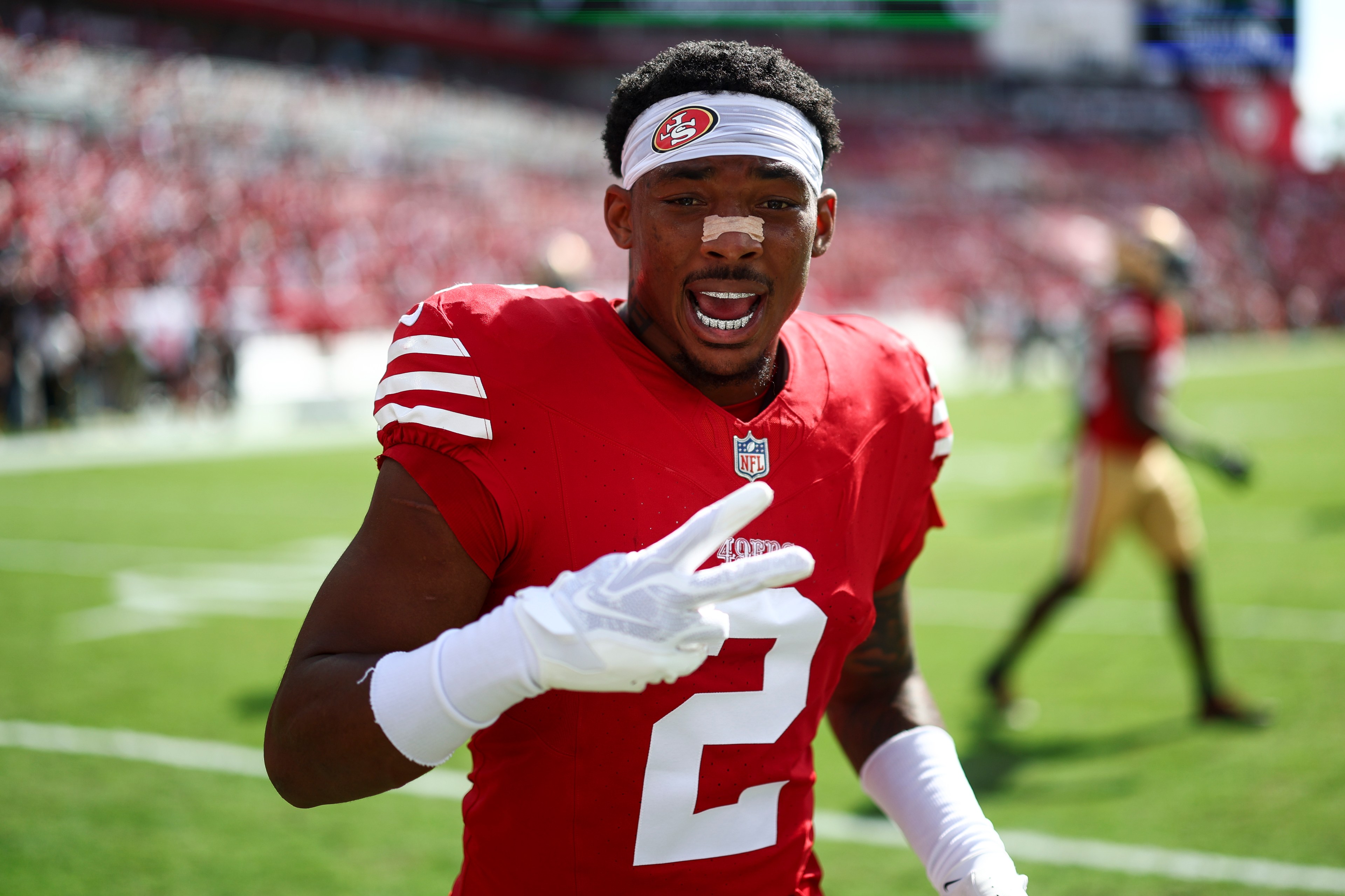 A football player in a red jersey, helmetless, gestures with two fingers. He's wearing a white headband and gloves, and stands on a sunny field.