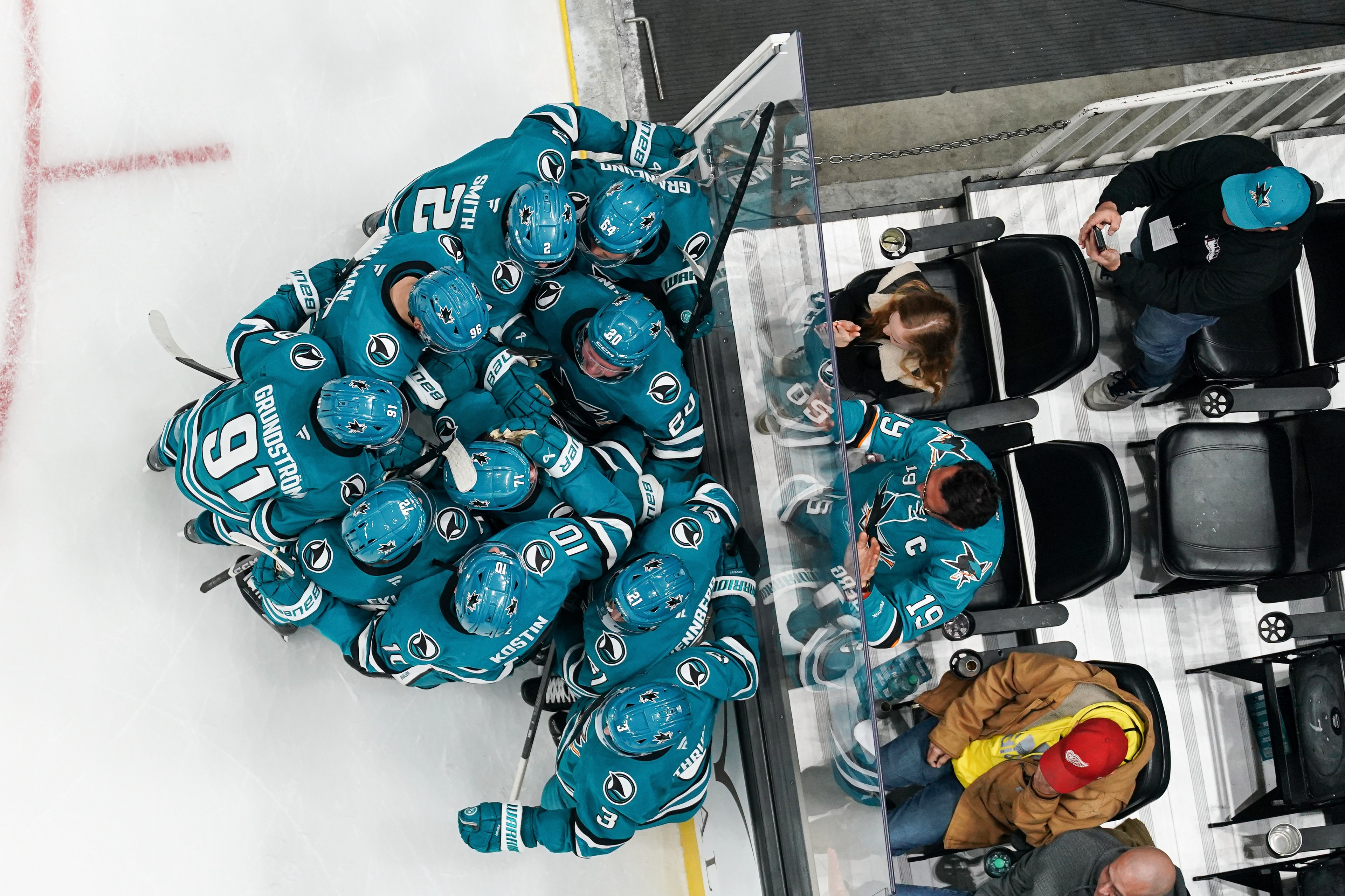 Hockey players in teal jerseys huddle near the ice, with fans seated behind glass. Some fans wear team jerseys, and one person is taking a photo.