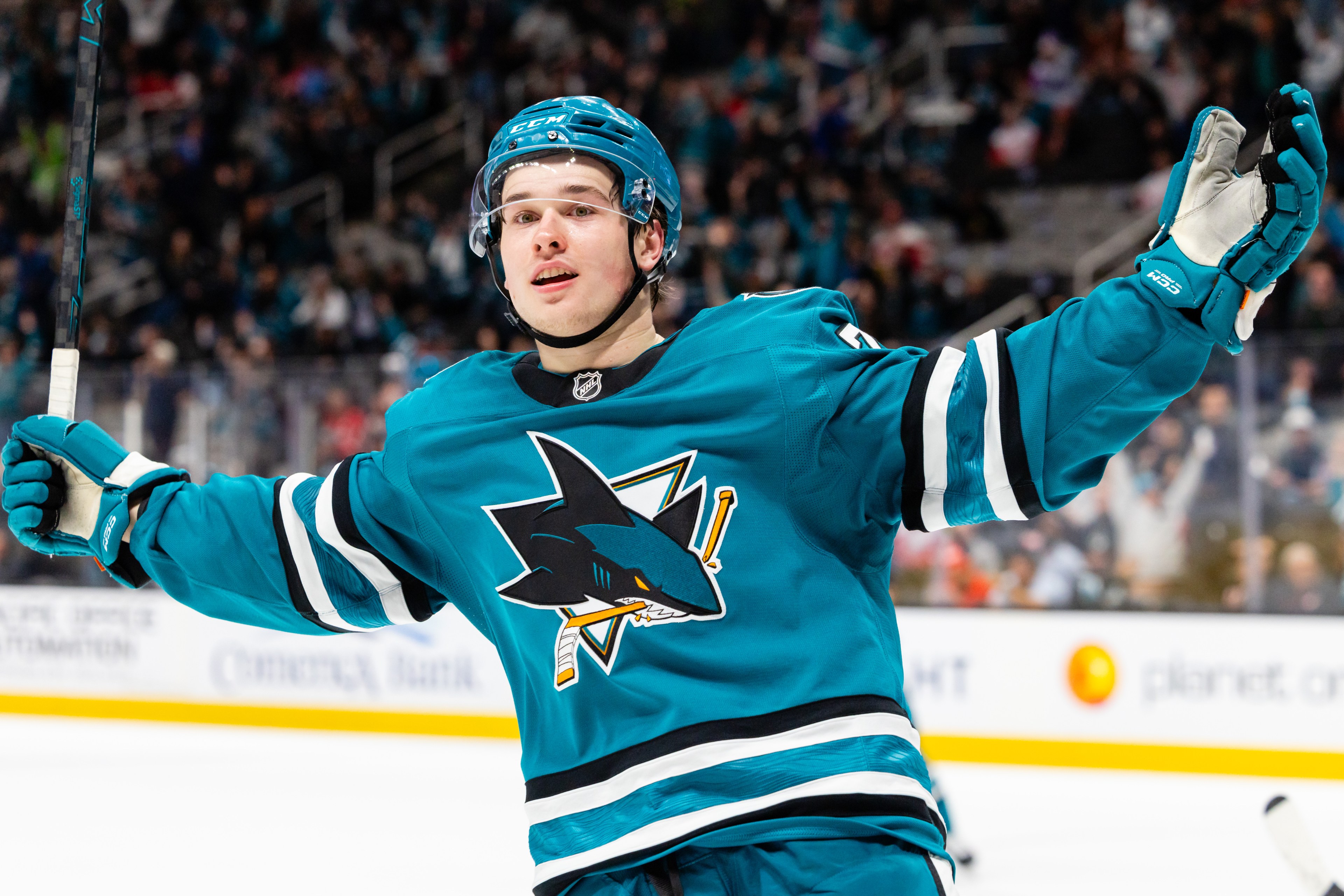 A hockey player in a teal jersey celebrates on the ice, arms wide, holding a hockey stick. The jersey features a shark logo. A crowd is blurred in the background.