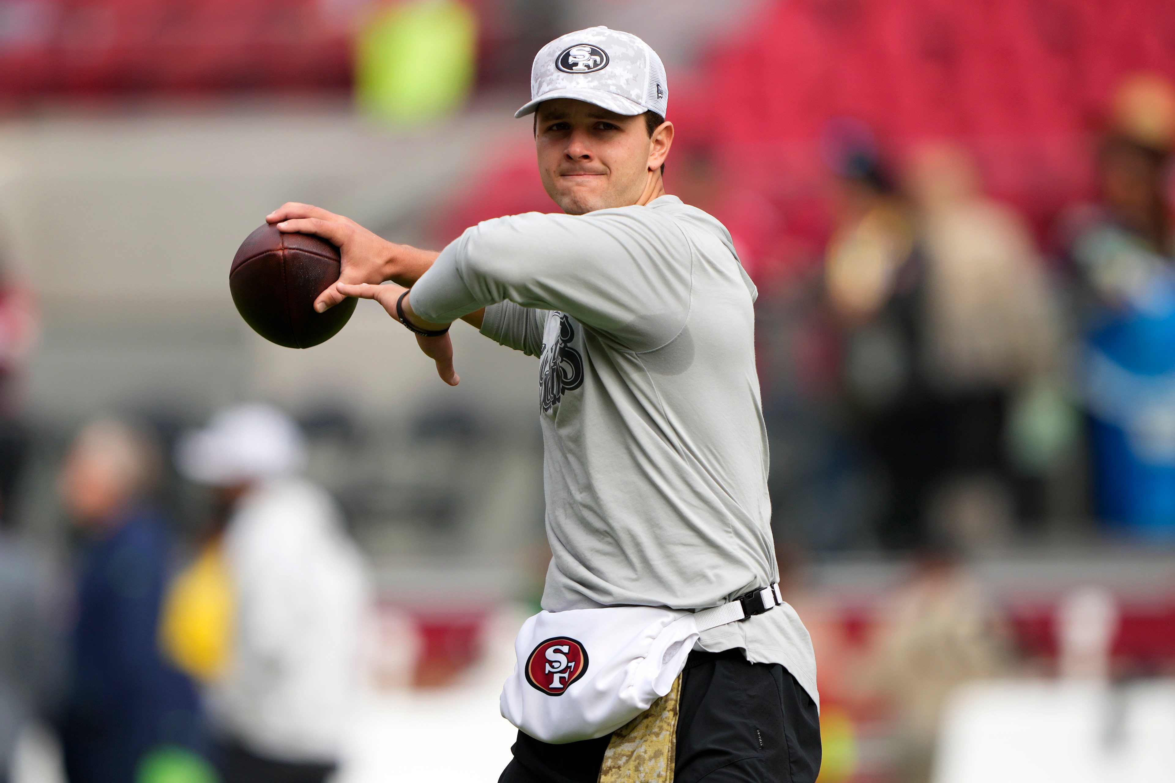 A person in a gray shirt and cap is preparing to throw a football. They wear a white pouch with a red and black logo. The background is a blurred stadium.