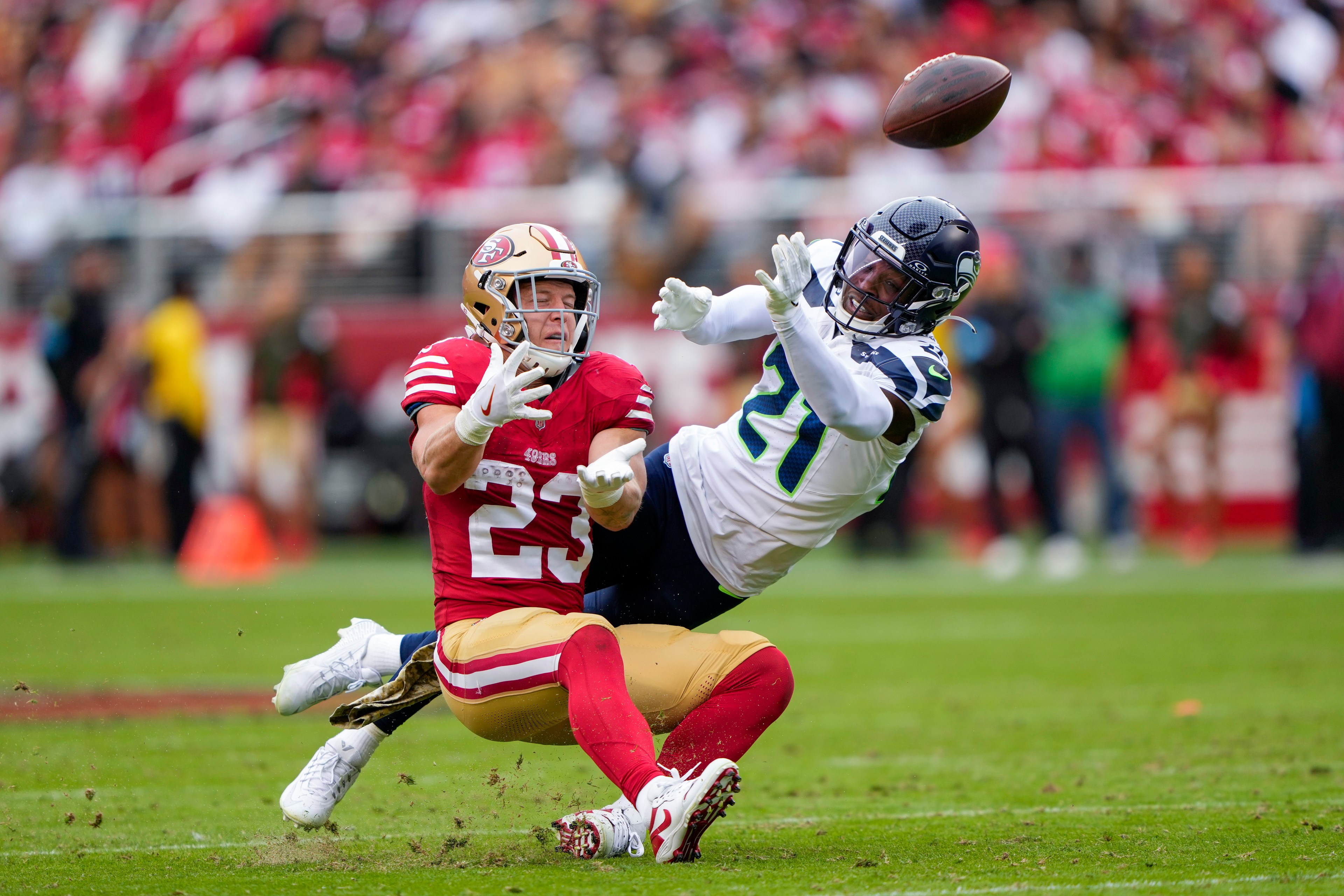 Two football players are in action; one in a red uniform is falling as he reaches for the ball, while the other in white tries to intercept it. The background is blurred.