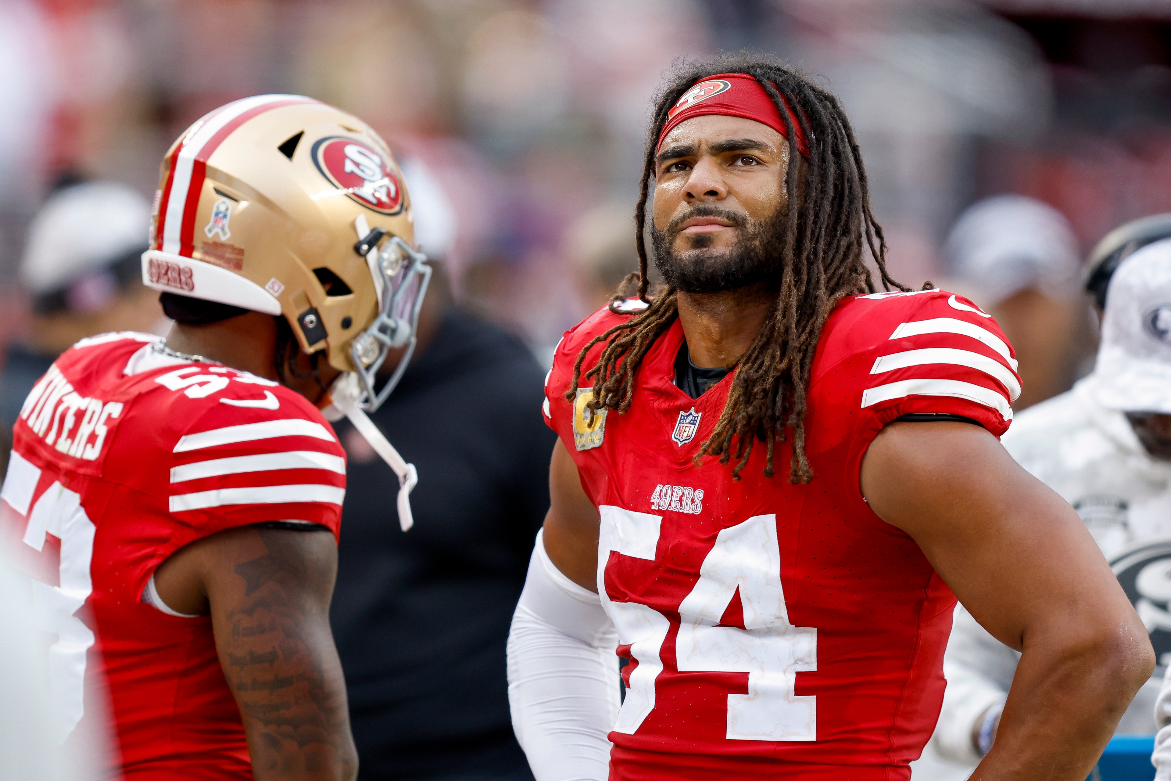 Two American football players in red San Francisco 49ers jerseys stand on the field. One wears a helmet, and the other has long hair and a headband.