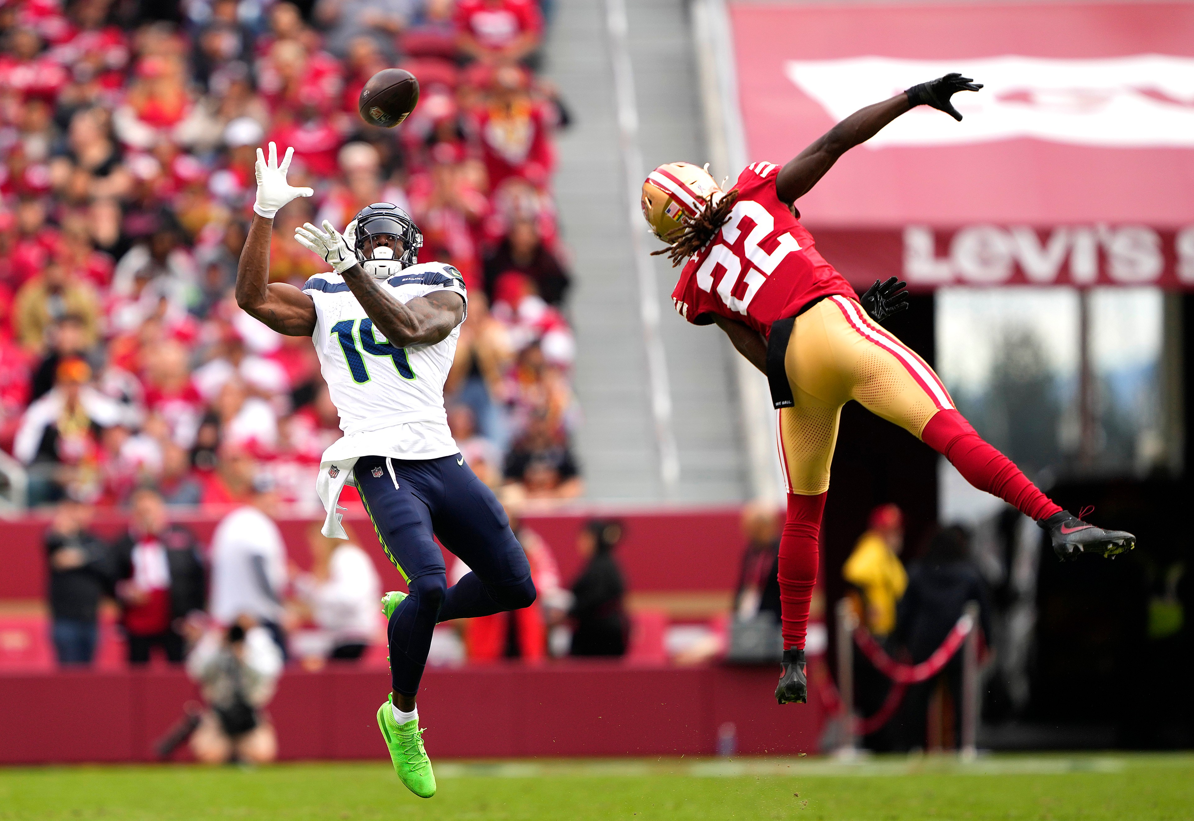 Two football players are jumping for a pass; one in a white and blue uniform with number 14, and the other in red and gold with number 22. The crowd is in the background.