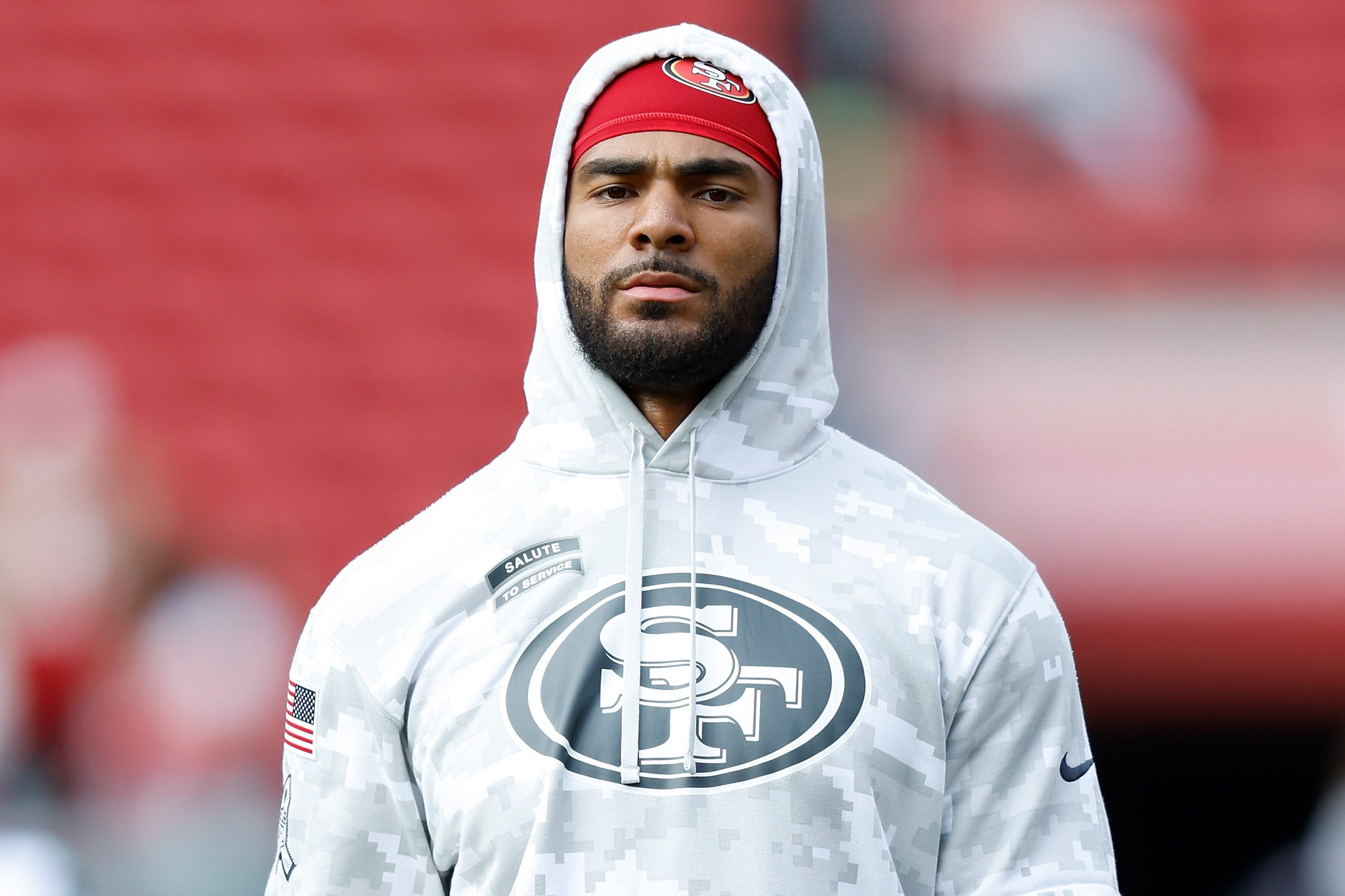 A man in a camouflage hoodie with an "SF" logo holds a football in a stadium. He wears a red cap, white gloves, and black shorts.