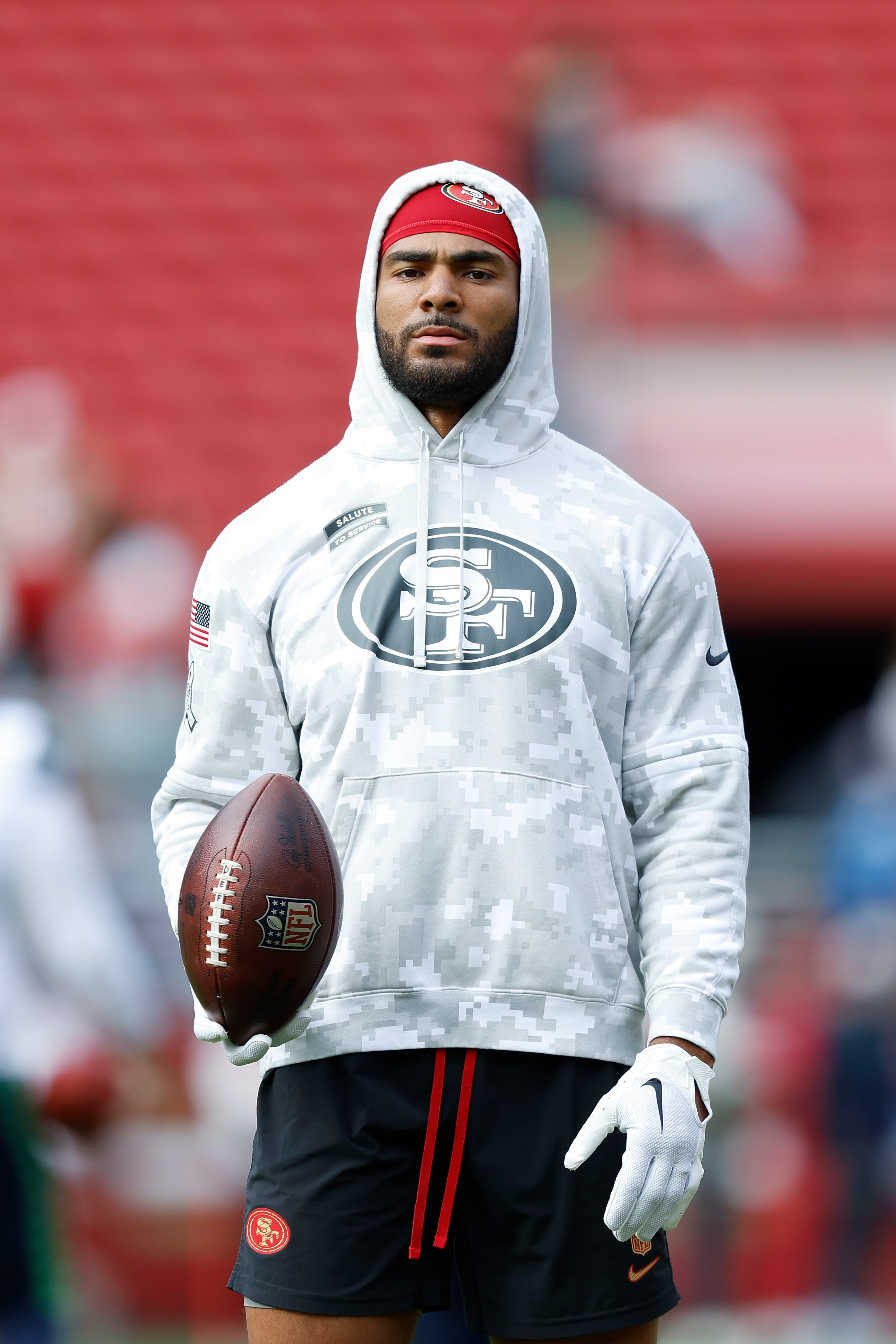 A man in a camouflage hoodie with an "SF" logo holds a football in a stadium. He wears a red cap, white gloves, and black shorts.