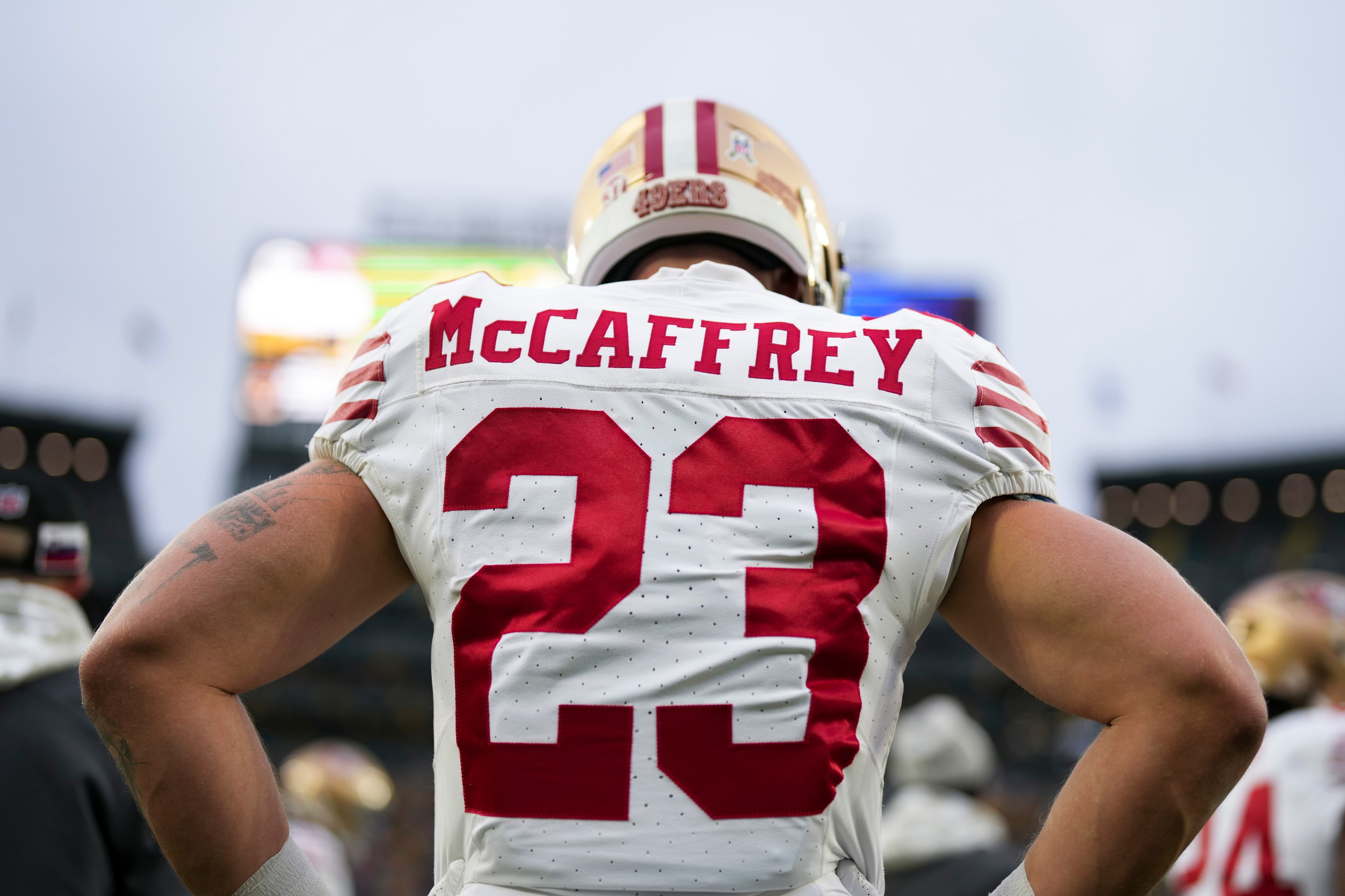 A football player wearing a white jersey with red stripes stands with his back to the camera. The name "McCaffrey" and the number "23" are visible, with a stadium in the background.