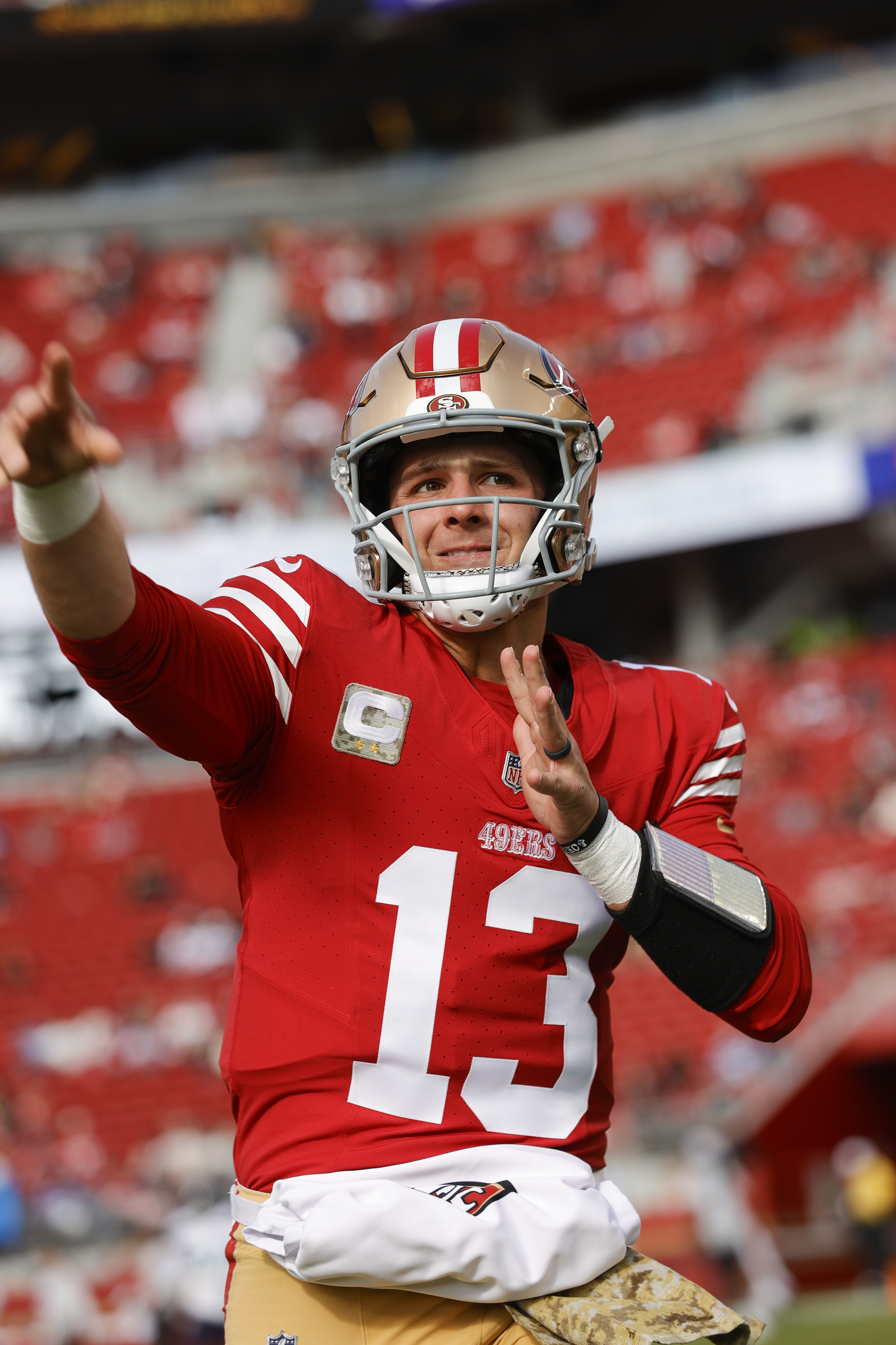 A football player in a red jersey and helmet is gesturing with his arm extended. The stadium background is blurred with red seats visible.