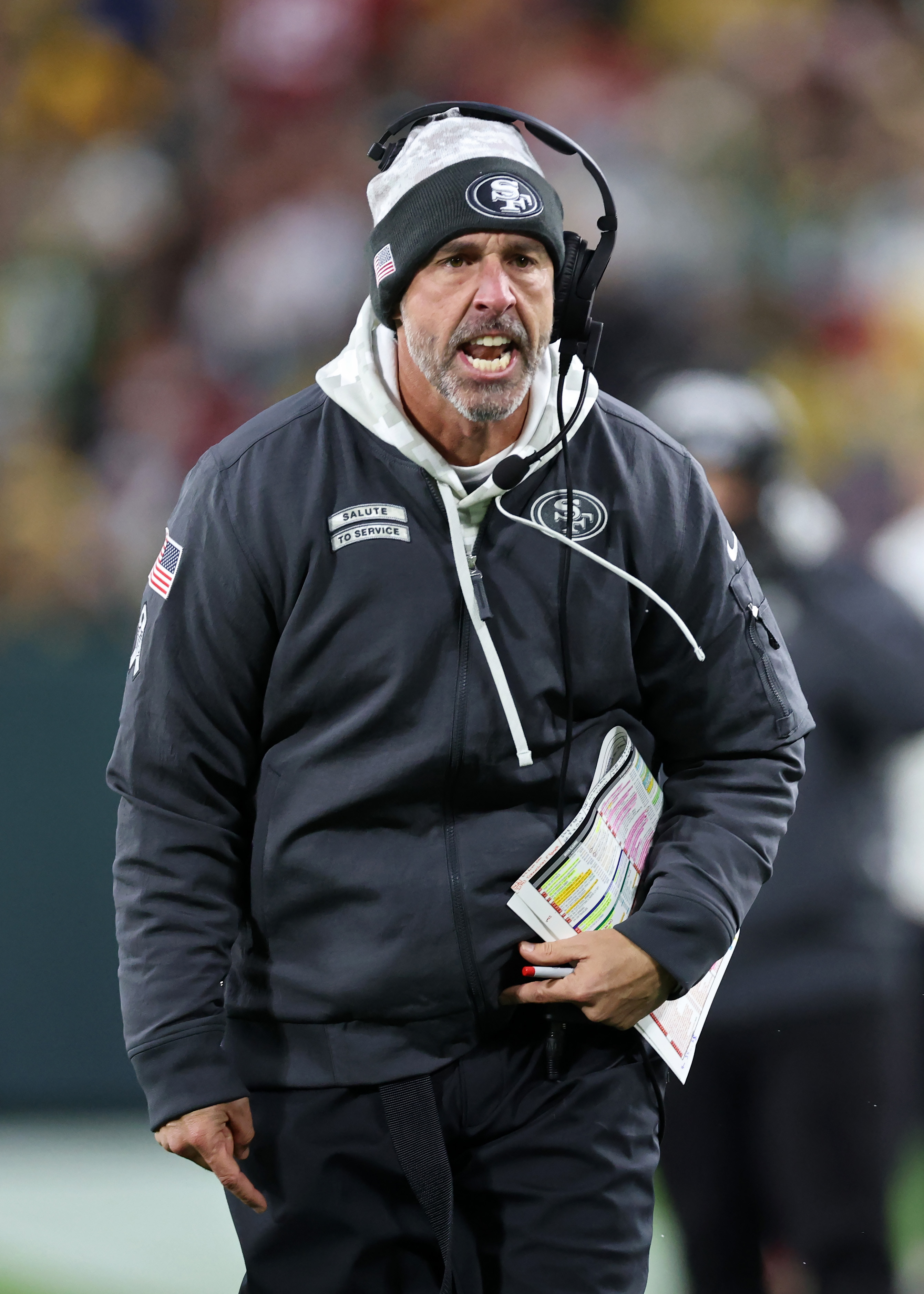 A coach wearing a beanie and headset is on a sports field, holding a folded playbook. He looks intense, with an open mouth, and wears a dark jacket.