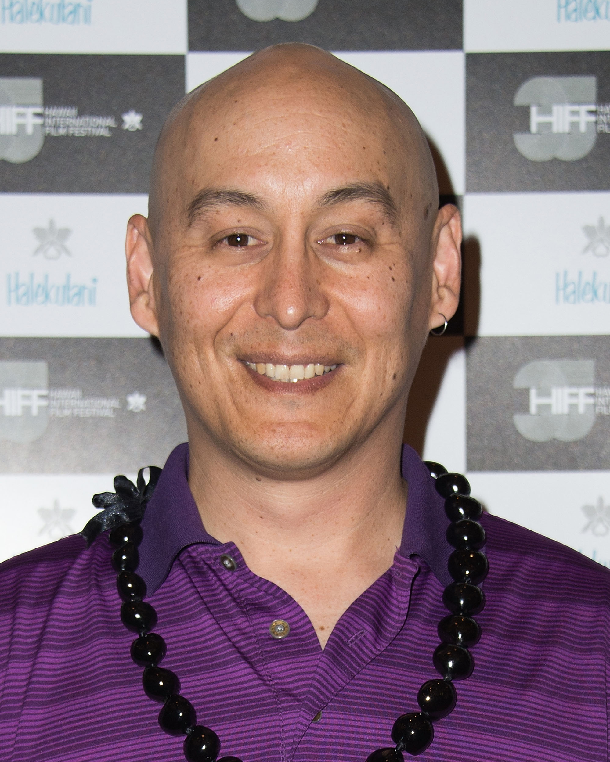 A smiling bald person is wearing a purple shirt and a black beaded necklace. The background features a checkered pattern with &quot;HIFF&quot; logos.