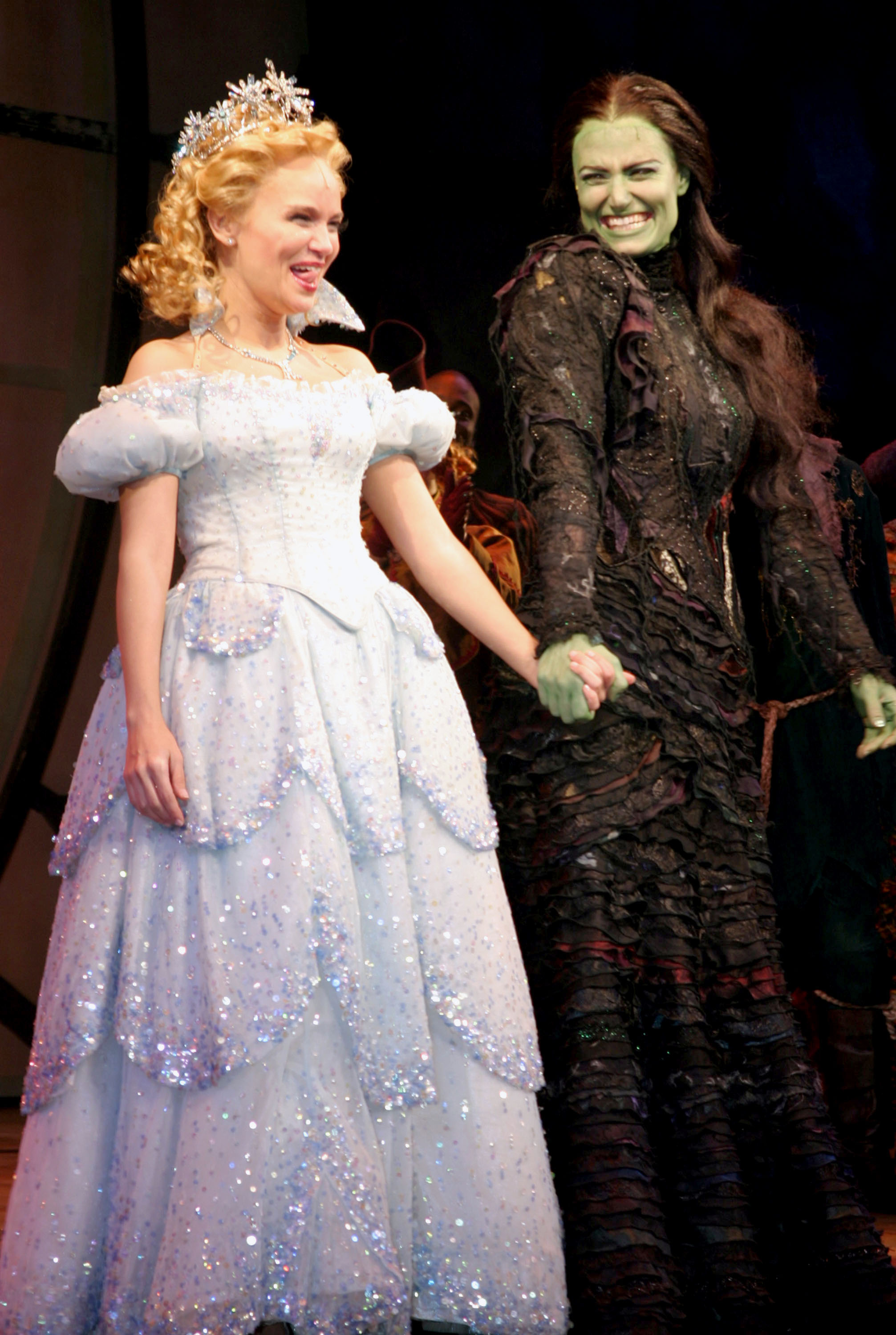 Two women are on stage holding hands; one in a sparkling blue gown with a tiara, and the other in a dark dress with green skin, both smiling.