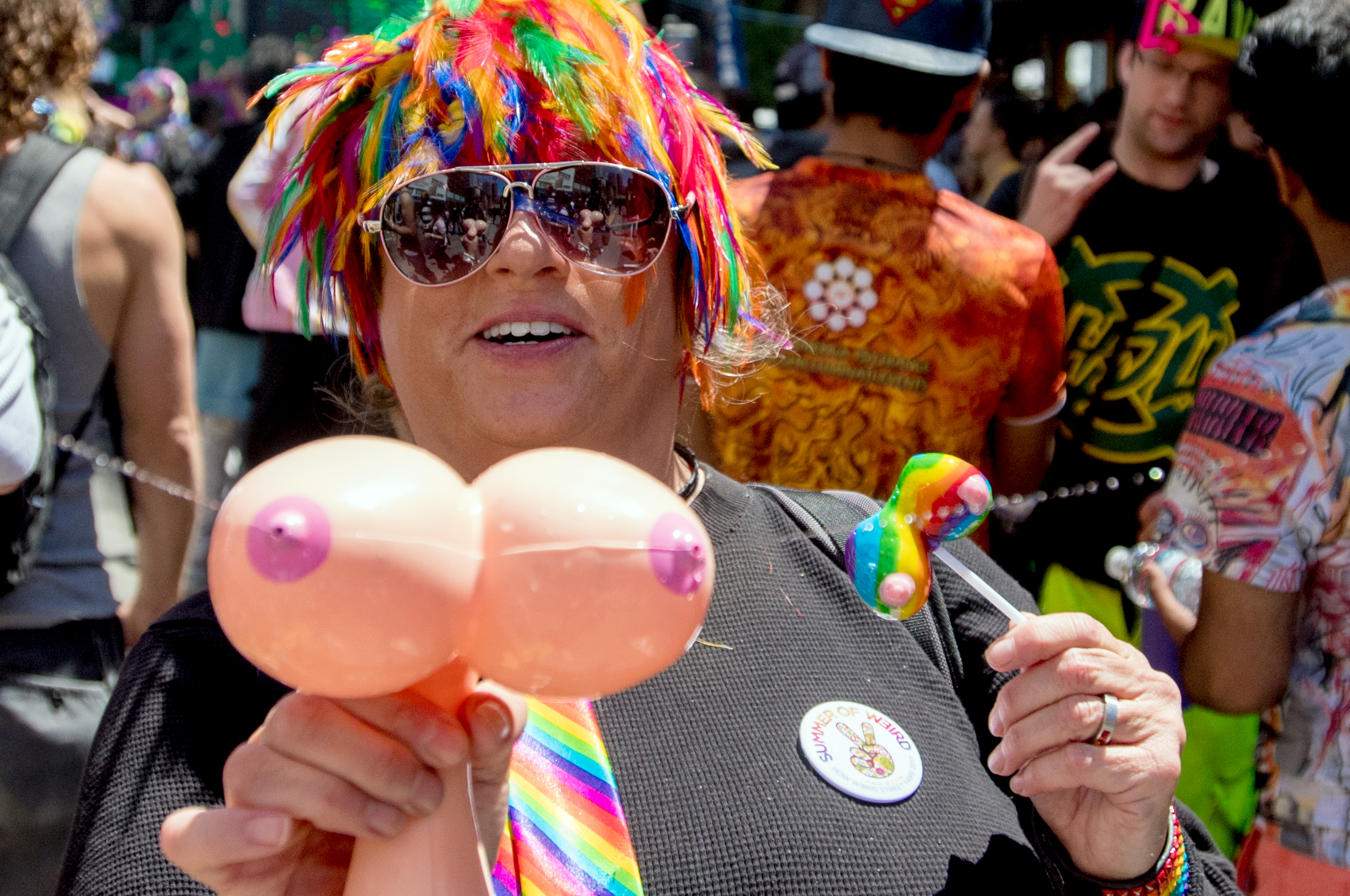 A person wearing a colorful wig and sunglasses holds a balloon sculpture and a rainbow-colored lollipop. They are in a lively crowd, wearing a rainbow tie.