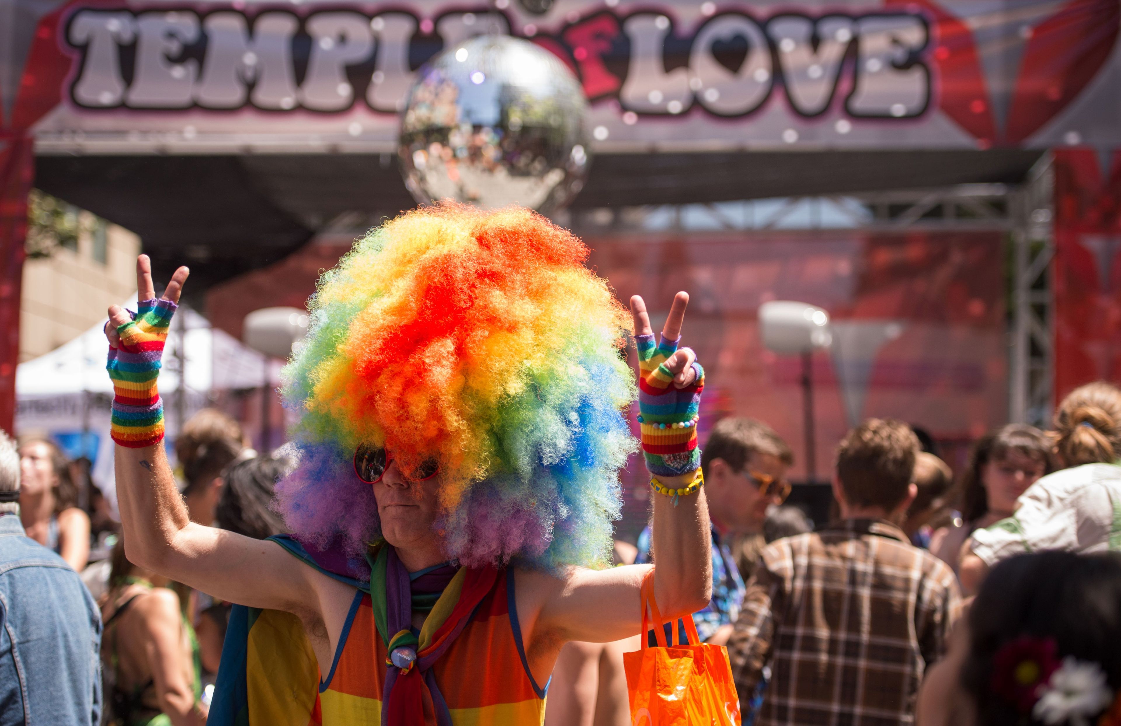 A person with a large rainbow afro, rainbow fingerless gloves, and sunglasses flashes peace signs at a lively outdoor event with a &quot;Temple of Love&quot; sign.