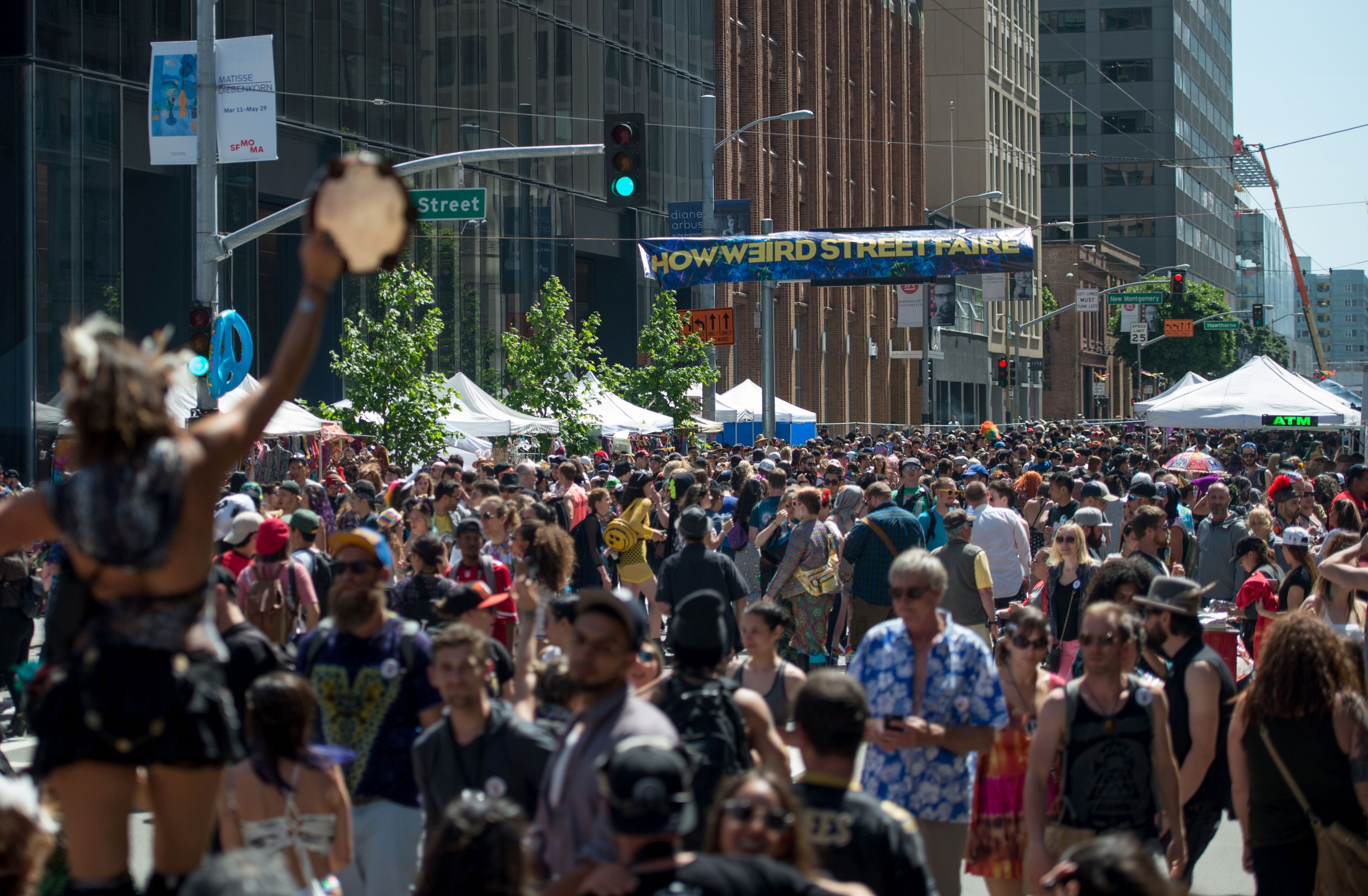 A large crowd fills a street during the How Weird Street Faire, with people in colorful outfits and a festive atmosphere. Tents and signs are visible in the background.