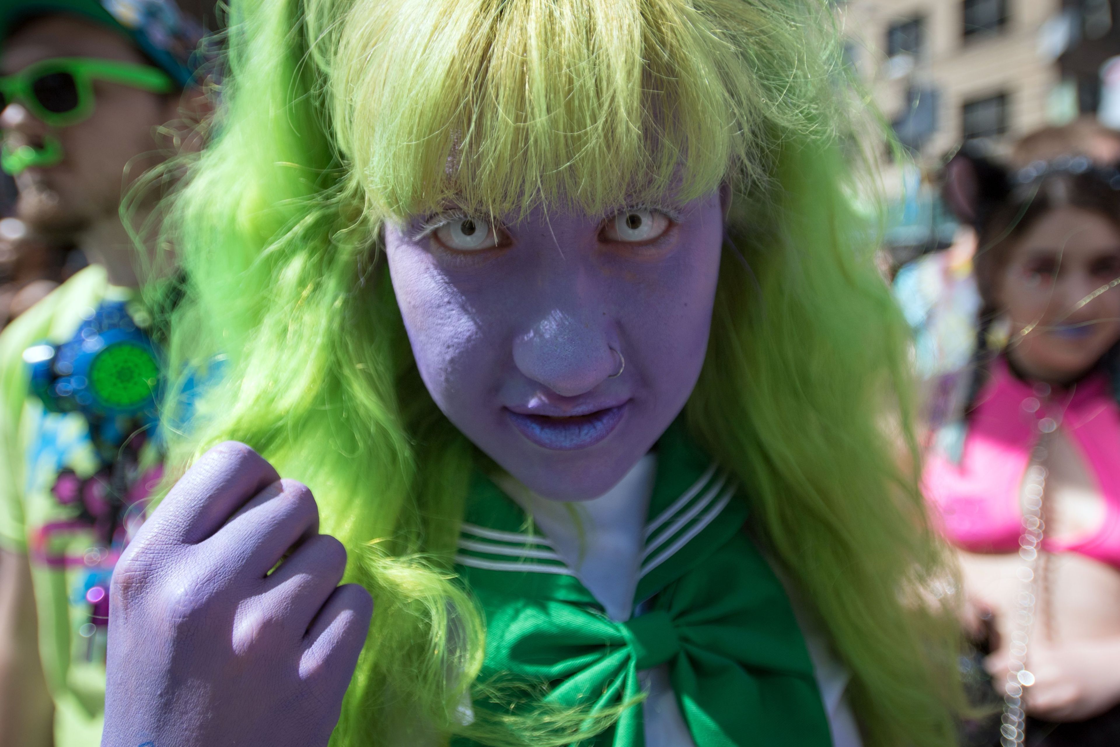 A person with bright green hair and purple skin wears a green tie and white outfit. Others dressed colorfully are blurred in the background.