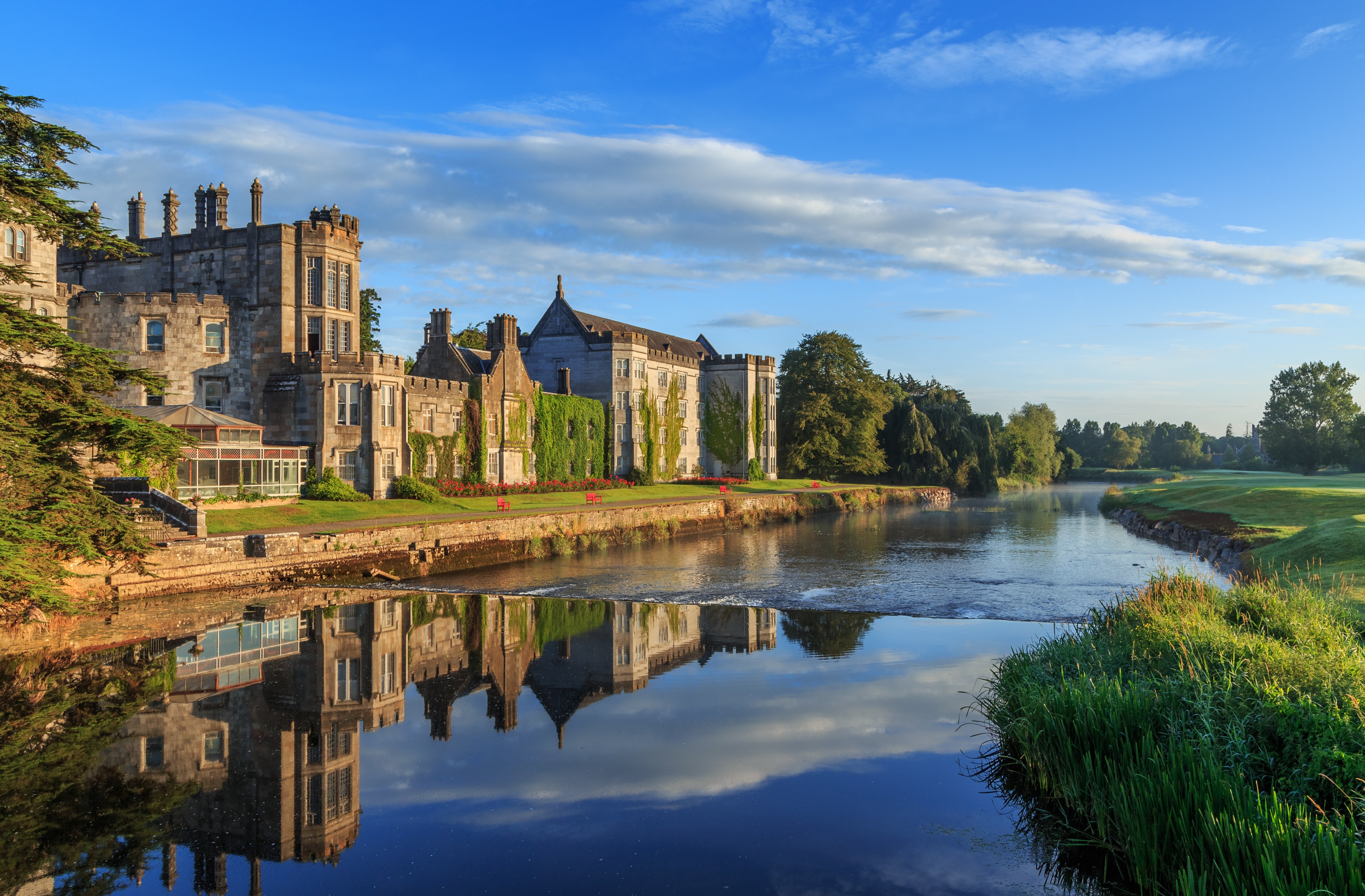 A grand, ivy-covered stone castle stands beside a calm river reflecting its image. The scene is surrounded by lush greenery under a clear blue sky.