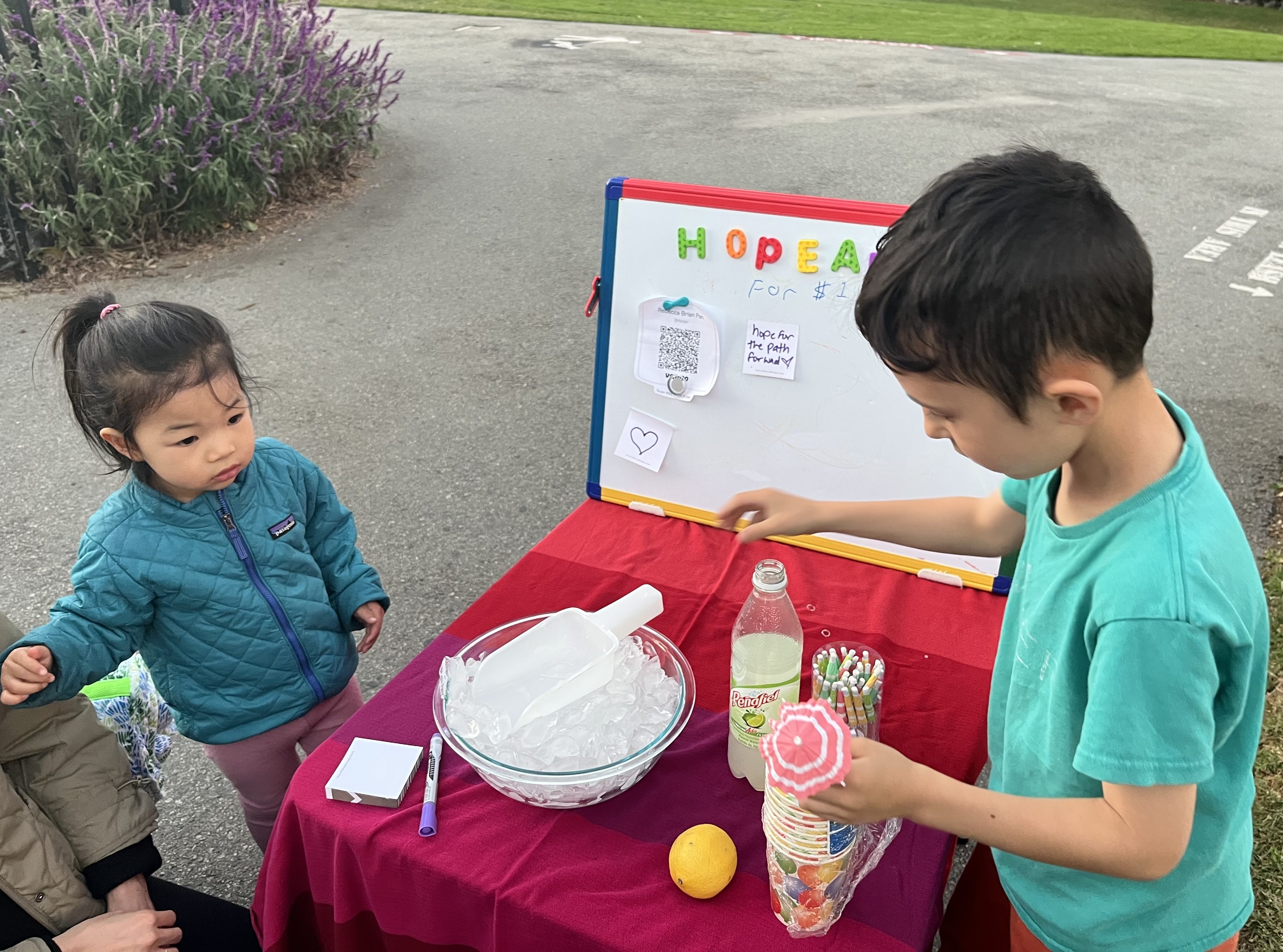 Two children are at a lemonade stand. One is organizing cups, and the other is looking on. There's a bowl of ice, a lemon, and a colorful sign on a board.